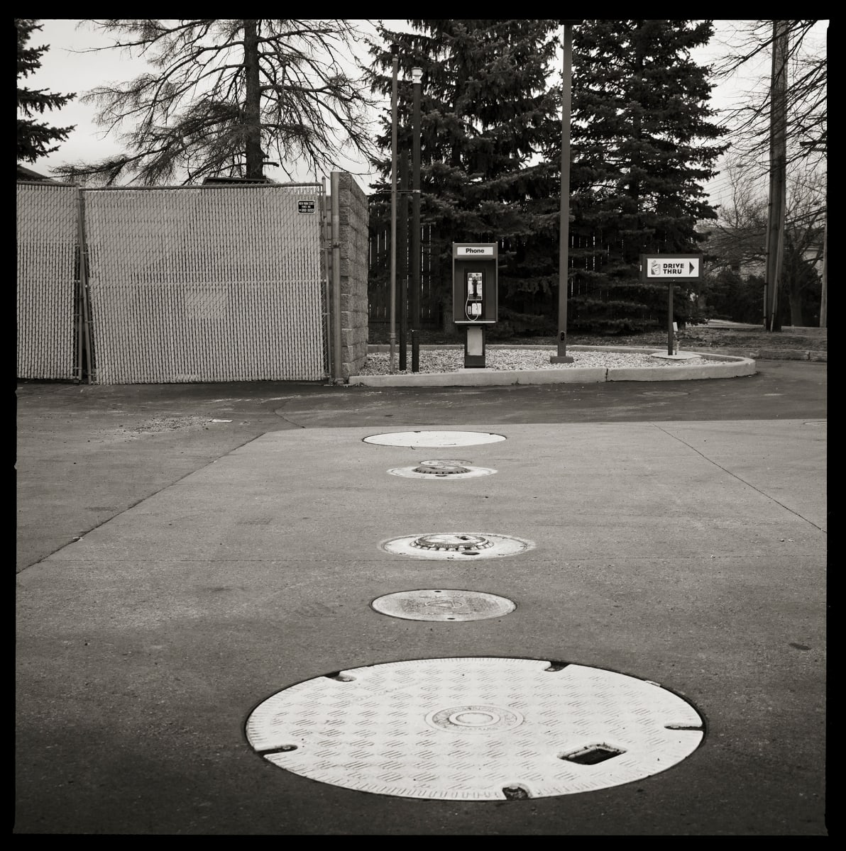 585.334.9884 – 3068 East Henrietta Road, Henrietta, NY 14467 by Eric T. Kunsman  Image: ID: A black and white image shows a payphone across the street.  In the street is a line of circular manhole covers that are white against the dark grey road.  Behind the payphone are trees.