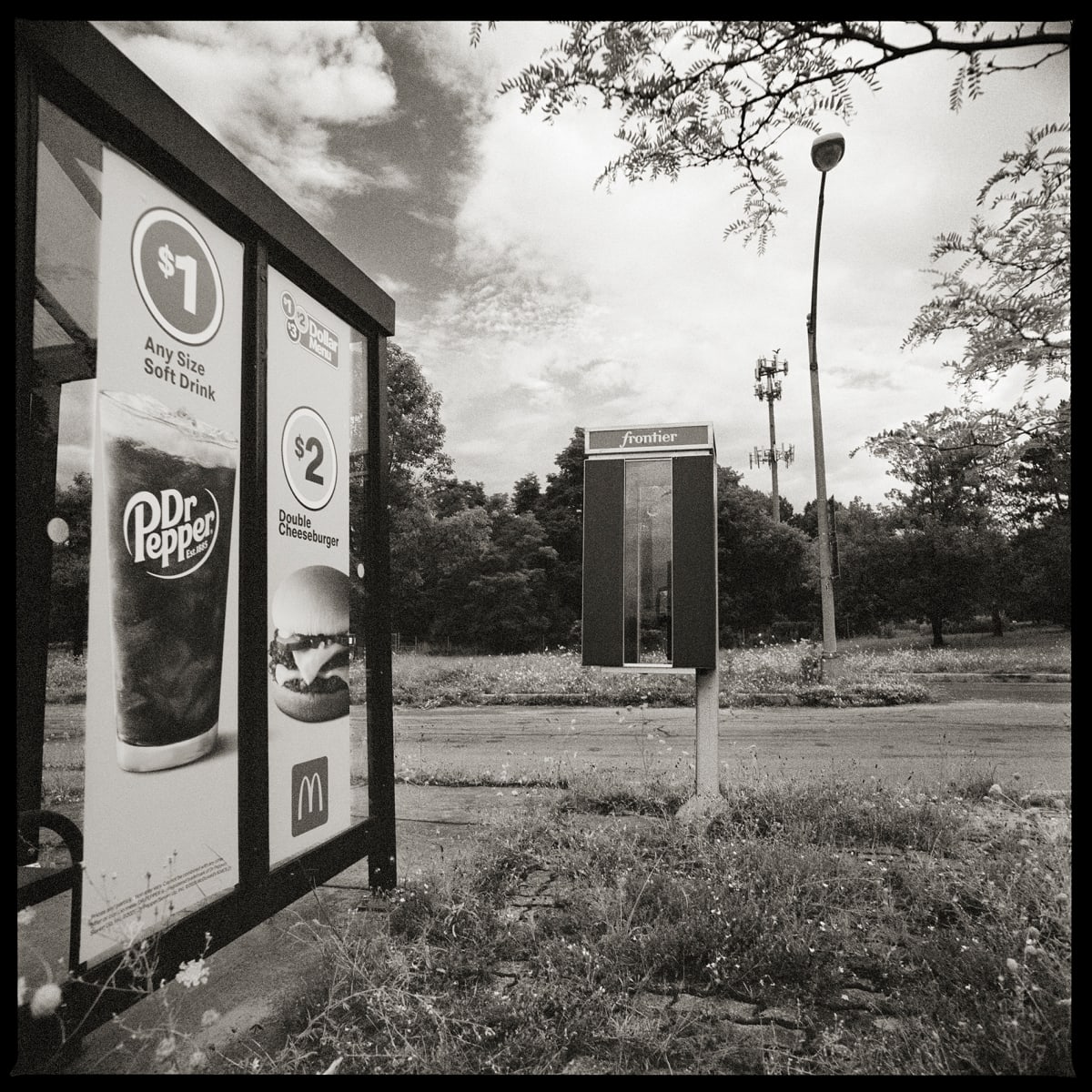 585.385.9292 – NYS Department of Transportation, 830 Pittsford-Victor Road, Pittsford, NY 14534 by Eric T. Kunsman  Image: ID: A black and white image shows a payphone standing to the right of a bus stop.  There is a road to the right of the payphone.