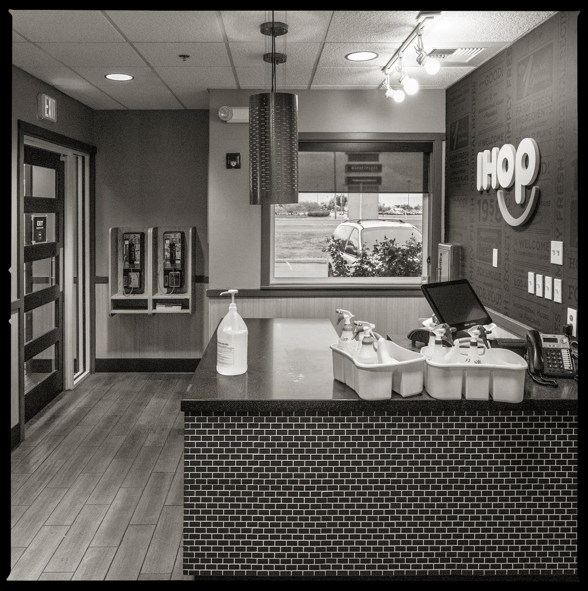 585.427.9524 & 585.427.9342 – IHOP, 556 Jefferson Road, Henrietta, NY, 14623 by Eric T. Kunsman  Image: ID: A black and white image shows an IHOP counter.  To the left, in the back there are two pay phones mounted on the wall.  To the right, there is a large white IHOP sign behind the counter on the wall.