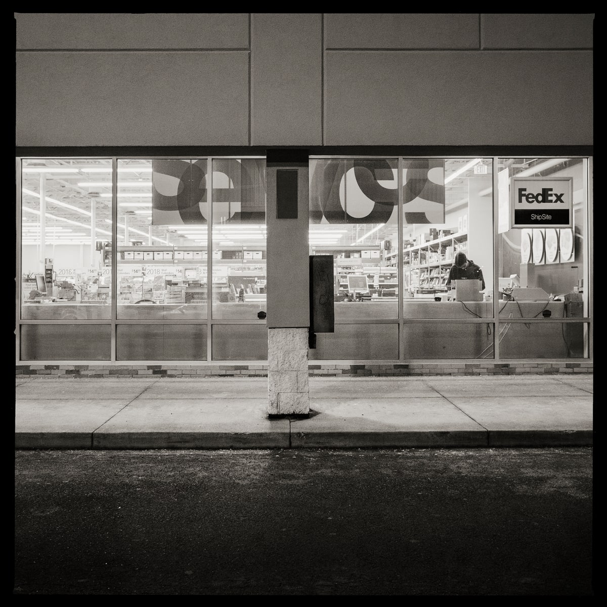 585.427.9694 – 3333 West Henrietta Road, Rochester, NY 14623 by Eric T. Kunsman  Image: ID: A black and white image shows a storefront with a pole in the center.  On the pole is a payphone.  There is a person visible through the store window.