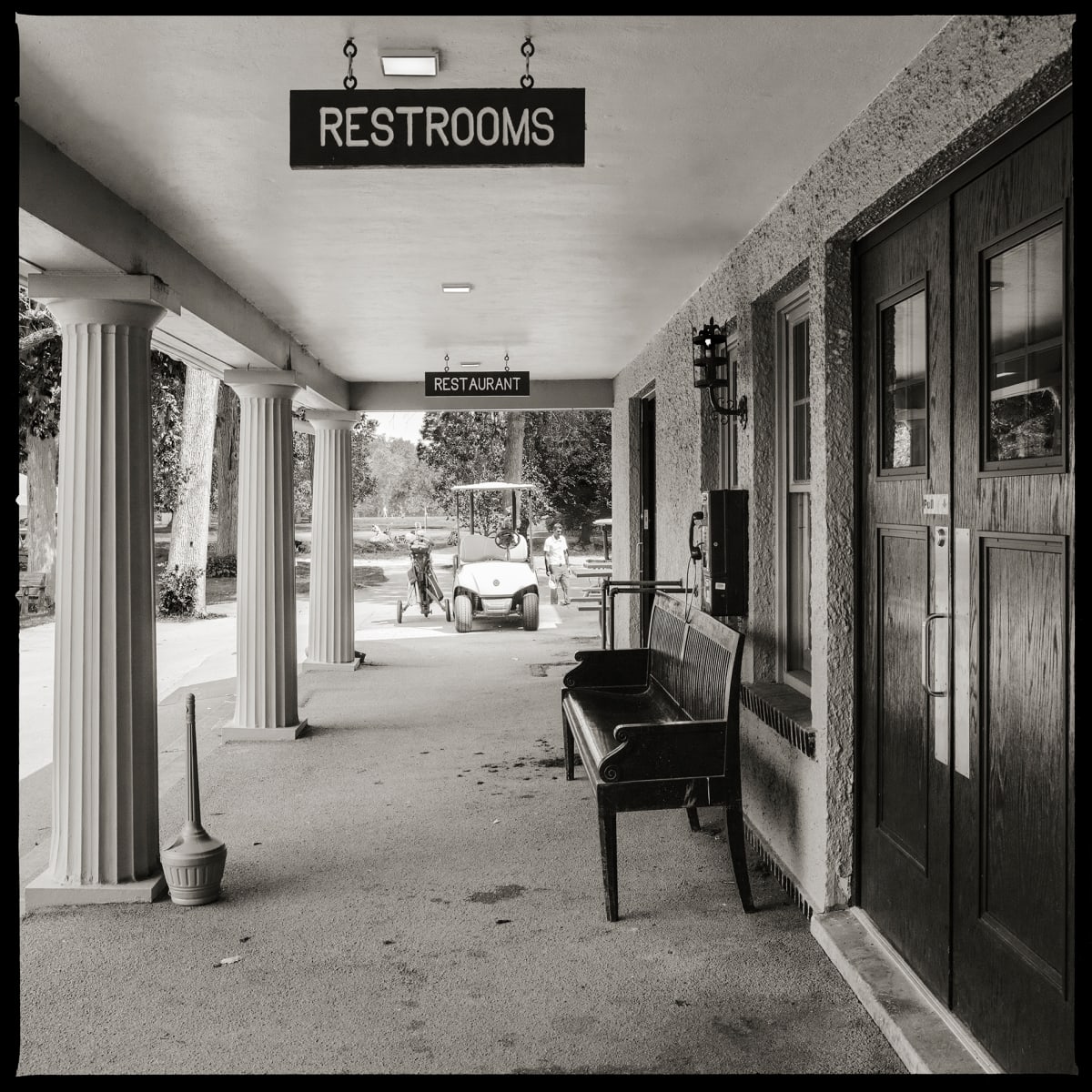 585.427.9724 – Genesee Valley Golf Course, 1000 East River Road, Rochester, NY 14623 by Eric T. Kunsman  Image: ID: A black and white image shows a pillared entrance way.  There is a black metal bench on the right.  Behind the bench is a wall mounted payphone.  There is a golf cart visible at the end of the entrance way.  There are signs for "Restrooms" and "Restaurant" hanging from the ceiling.