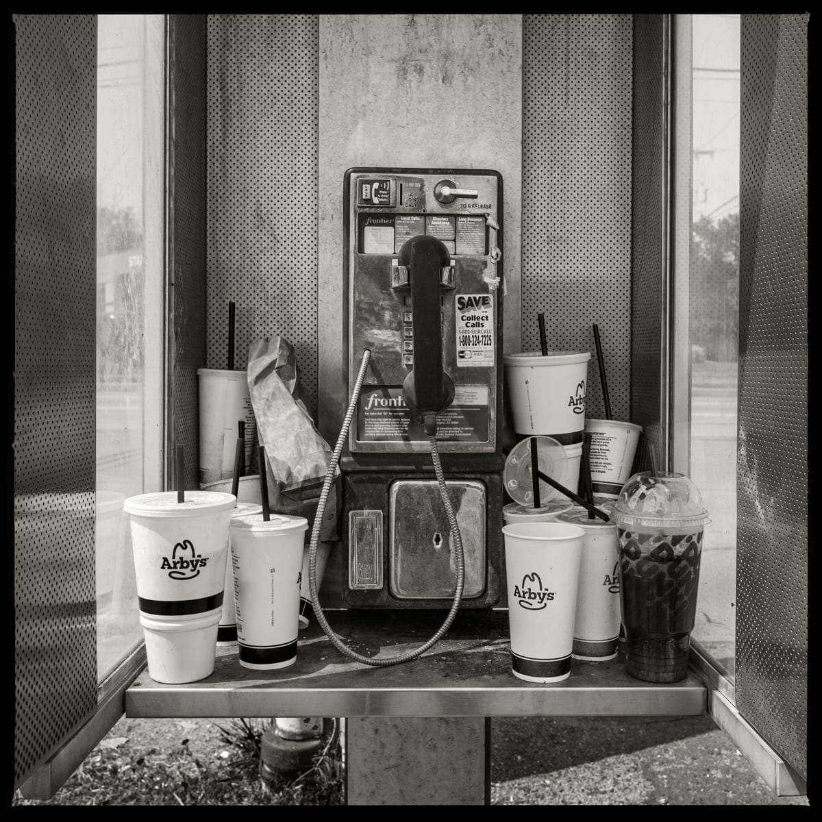 585.427.9848 – Stoney’s Plaza, 2852 West Henrietta Road, Henrietta, NY 14623 by Eric T. Kunsman  Image: ID: A black and white image shows a close up image of a payphone.  The phone is on the hook.  Used fast food cups surround the phone.