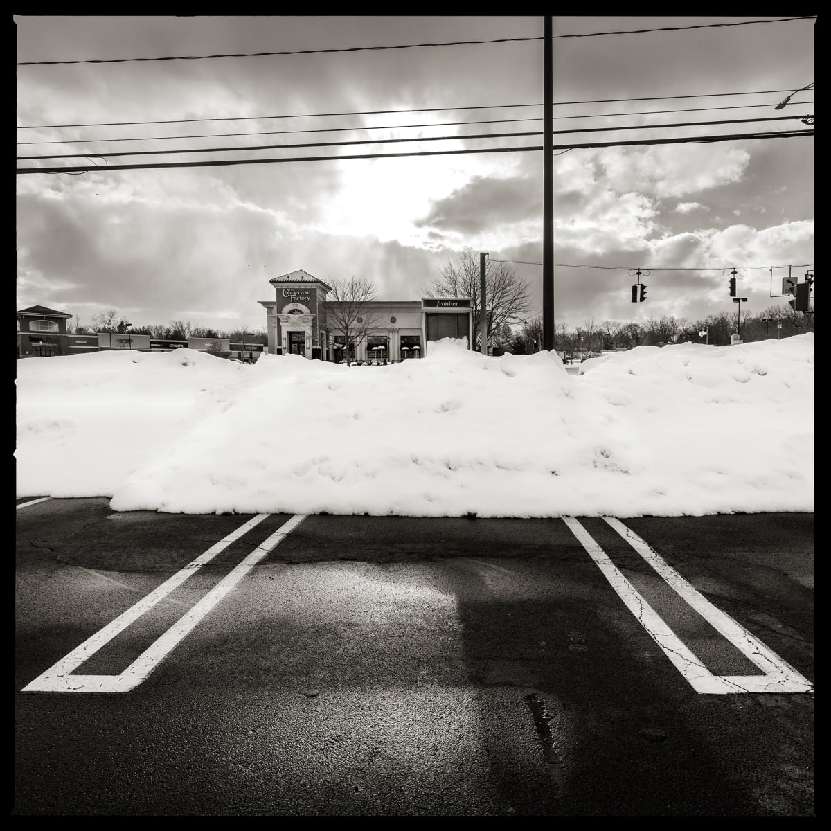 585.586.9201 – Pittsford Place Mall, 3300 Monroe Avenue, Rochester, NY 14618 by Eric T. Kunsman  Image: ID: A black and white image shows a payphone covered in snow.  The foreground is a parking lot and the background has a Cheesecake Factory.  The sky is cloudy with one patch of sun in the middle.  