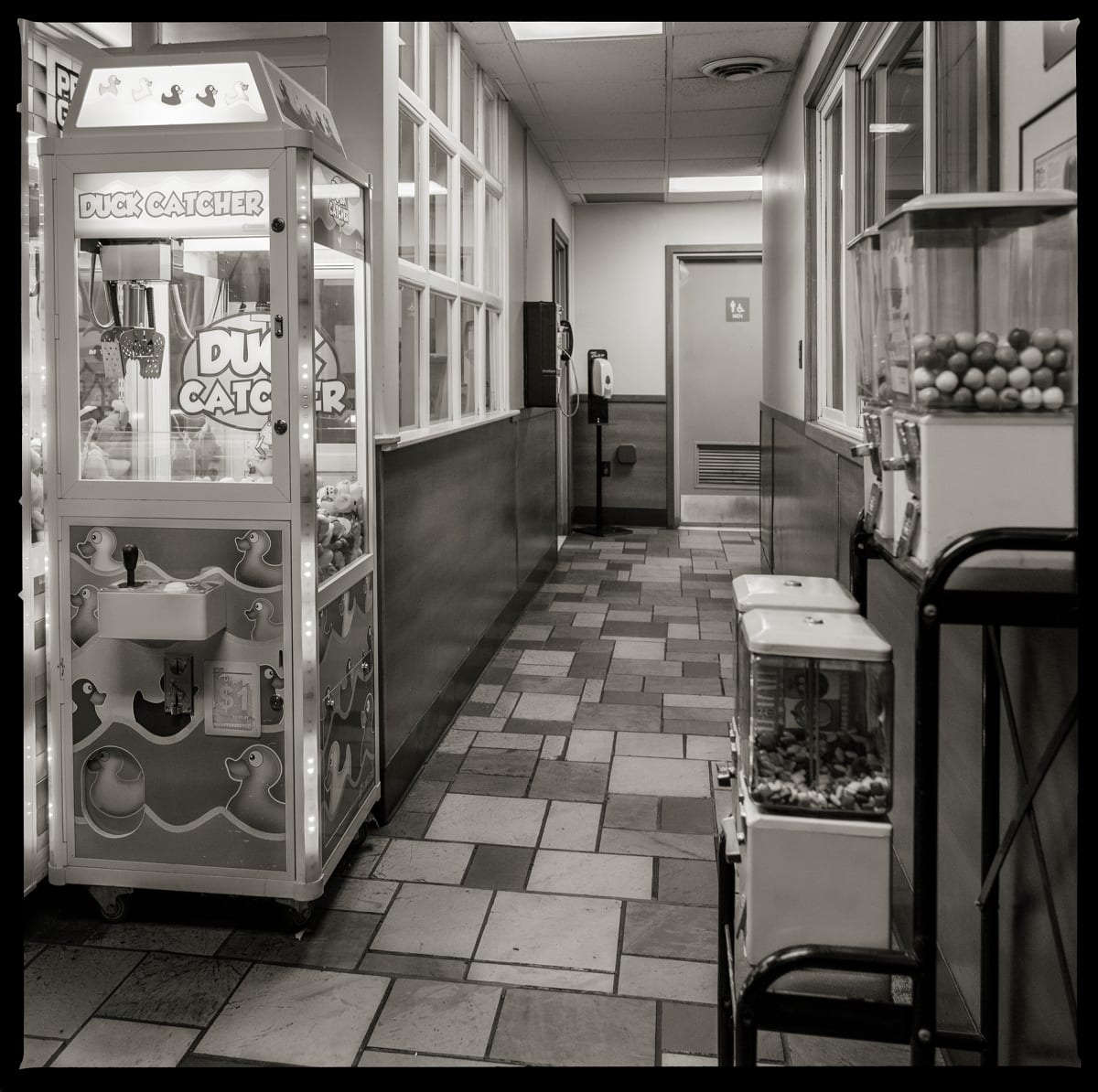 585.663.9688 – Schaller’s, 965 Edgemere Drive, Rochester, NY 14612 by Eric T. Kunsman  Image: ID: A black and white image shows a corridor with a payphone on the left wall.  There is a rubber duck claw machine on the left and gum ball machines on the right.  The floor is a patterned tile.