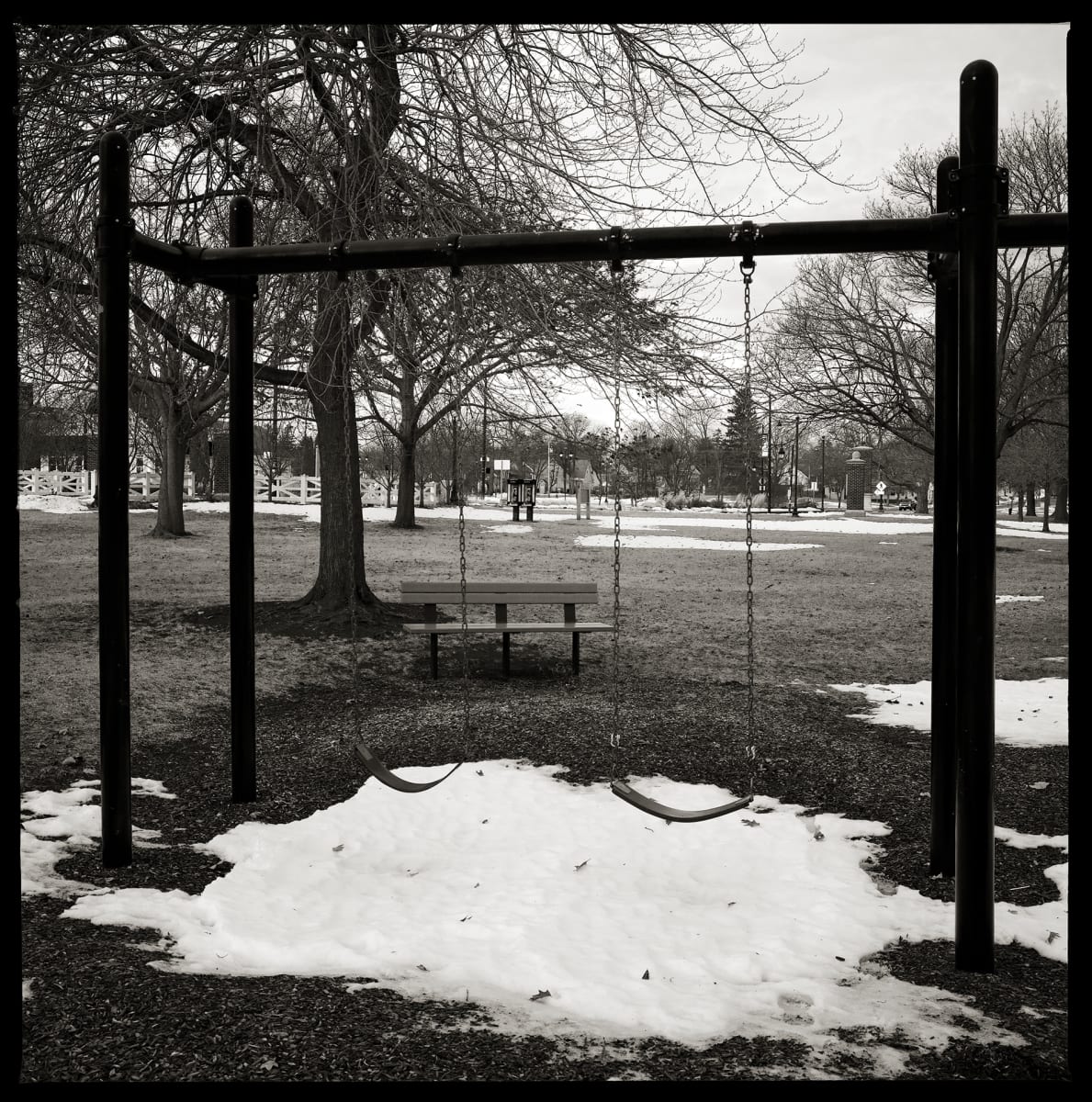 585.663.9803 & 585.663.9611 – 188 Beach Avenue, Rochester, NY 14612 by Eric T. Kunsman  Image: ID: A black and white image shows a swing set with two swings. There is a pile of snow beneath the swings.  There is a wooden bench and tree behind the left swing. In the distance, there is a pair of pay phones between the swings.