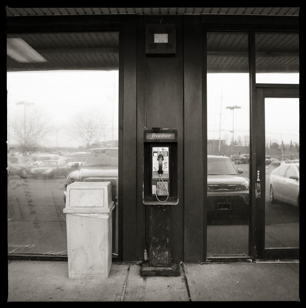585.671.9899 – Bay Center Plaza, 1217 Bay Road, Webster, NY 14580 by Eric T. Kunsman  Image: ID: A black and white image shows a payphone against a blank portion of wall that is surrounded by semi-reflective glass windows and doors.  There is a grey garbage can to the left of the payphone