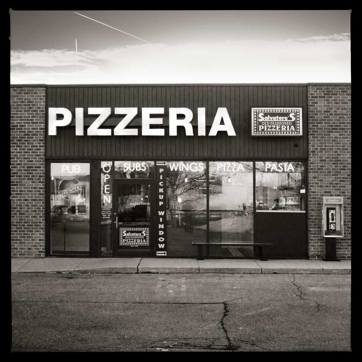 585.671.9950 – Salvatore’s Pizza, 1217 Bay Road, Webster, NY 14580 by Eric T. Kunsman  Image: ID: A black and white image shows a brick building that houses a Salvatore's Pizza.  There is a glass window and door advertising the pizzeria, with a big sign that says "PIZZERIA" above the door.  To the right of the windows is a payphone.