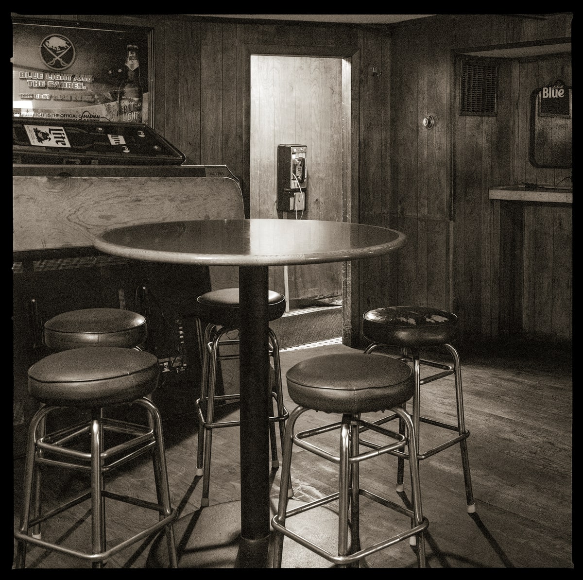585.872.9919 – Coach’s Sports Bar, 19 West Main Street, Webster, NY 14580 by Eric T. Kunsman  Image: ID: A black and white image shows a payphone in a back closet of a bar.  In front of the payphone, there is a circular table with five bar stools around it.  The loser light is on, making the payphone visible.