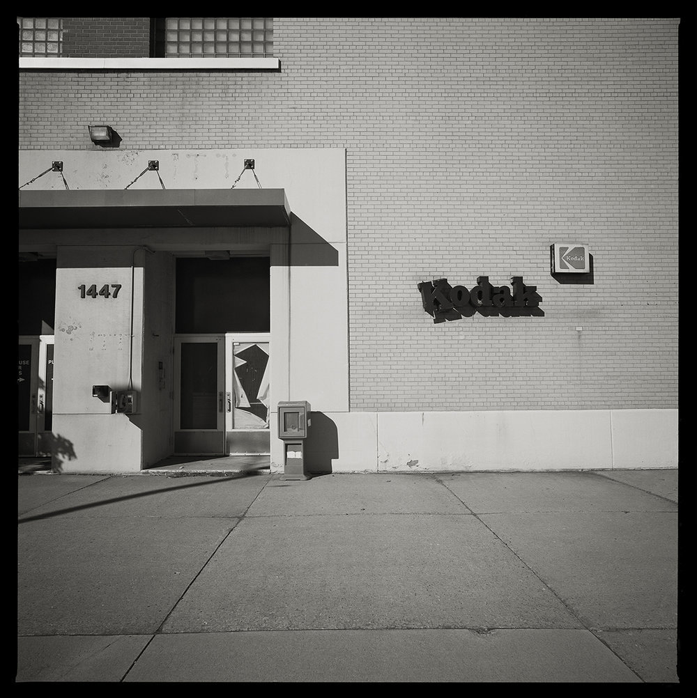 Kodak Hawkeye Building, Rochester, NY by Eric T. Kunsman  Image: ID: A black and white image shows a building entrance way on the left.  On the wall to the right of the entrance there are two Kodak signs.  One sign has a bubbly font and the other is the classic Kodak insignia.