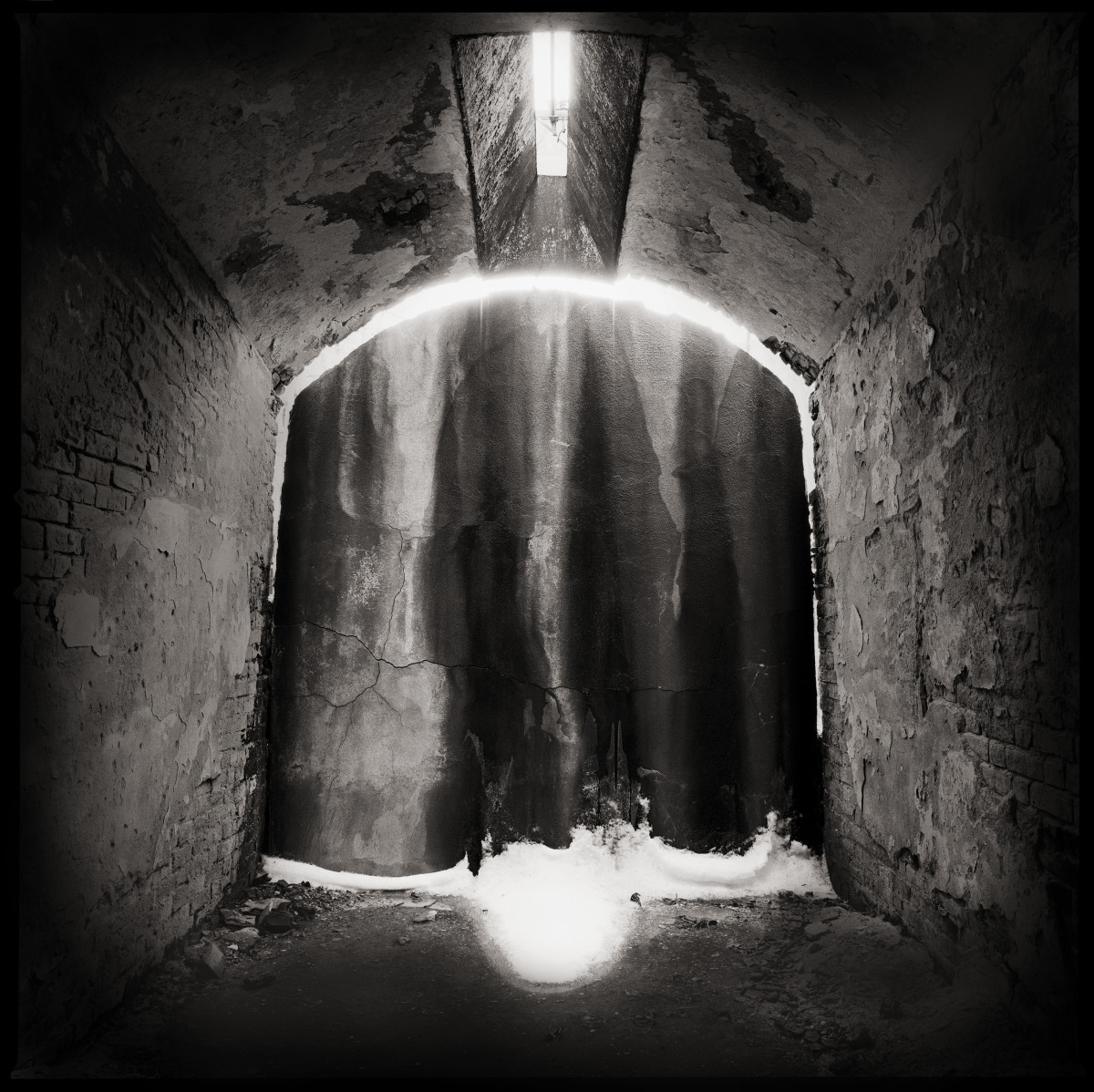 The Drapery by Eric T. Kunsman  Image: ID: A black and white image shows a corridor that ends with a drape.  The ceiling is curved and there is a window in the ceiling.  The light from the window highlights the dust in the air.  There is light beyond the drape which is seen at the bottom and top of the drape.