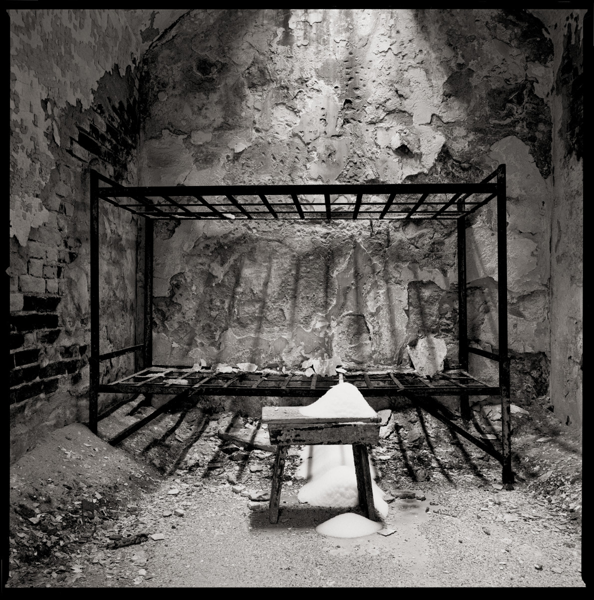Ashes by Eric T. Kunsman  Image: ID: A black and white image shows a cell room with a bunkbed frame against the back wall.  In front of the bed frame there is a wooden desk that has a pile of ashes on it as well as underneath it.  There are piles of dust and debris on the floor and the plaster has cracked off the brick walls.