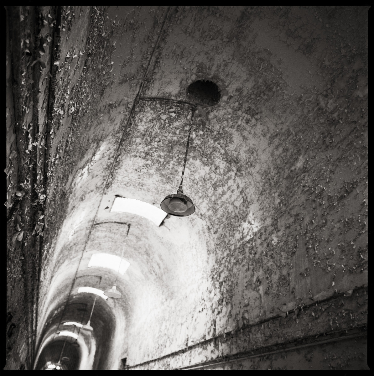 Infirmary Wing, West by Eric T. Kunsman  Image: ID: A black and white image shows tilted corridor with a curved roof.  There is light coming from the end of the corridor, but the end is not fully visible due to the tilted angle.  There are hanging light fixtures from the ceiling.