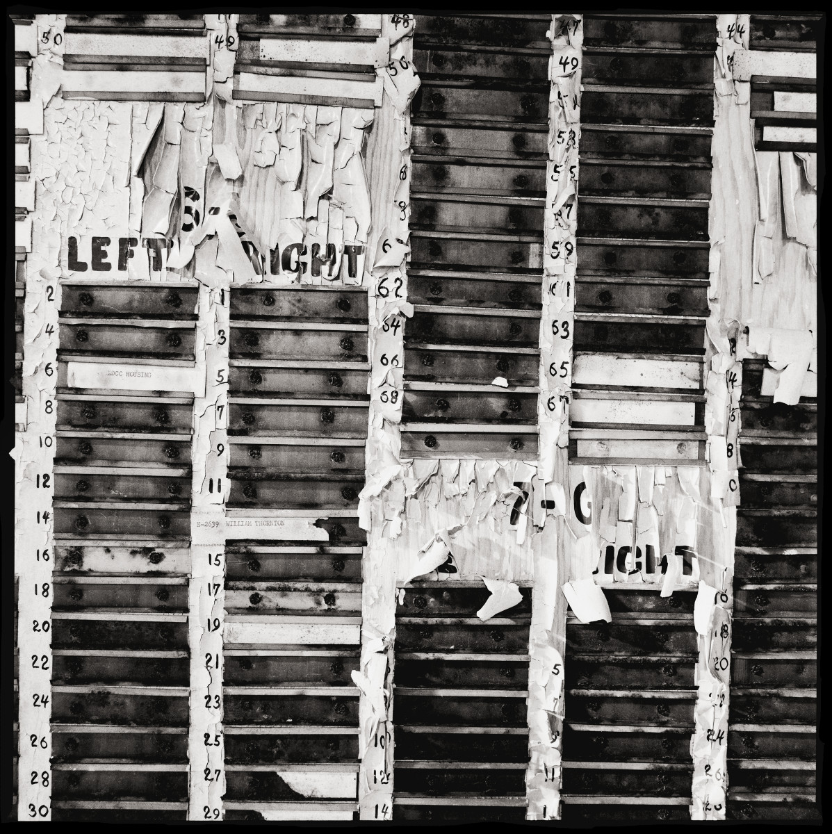 Control Board by Eric T. Kunsman  Image: ID: A black and white image shows what is left of the control board.  The slots are empty and the paint is peeling.  The slots are each numbered.