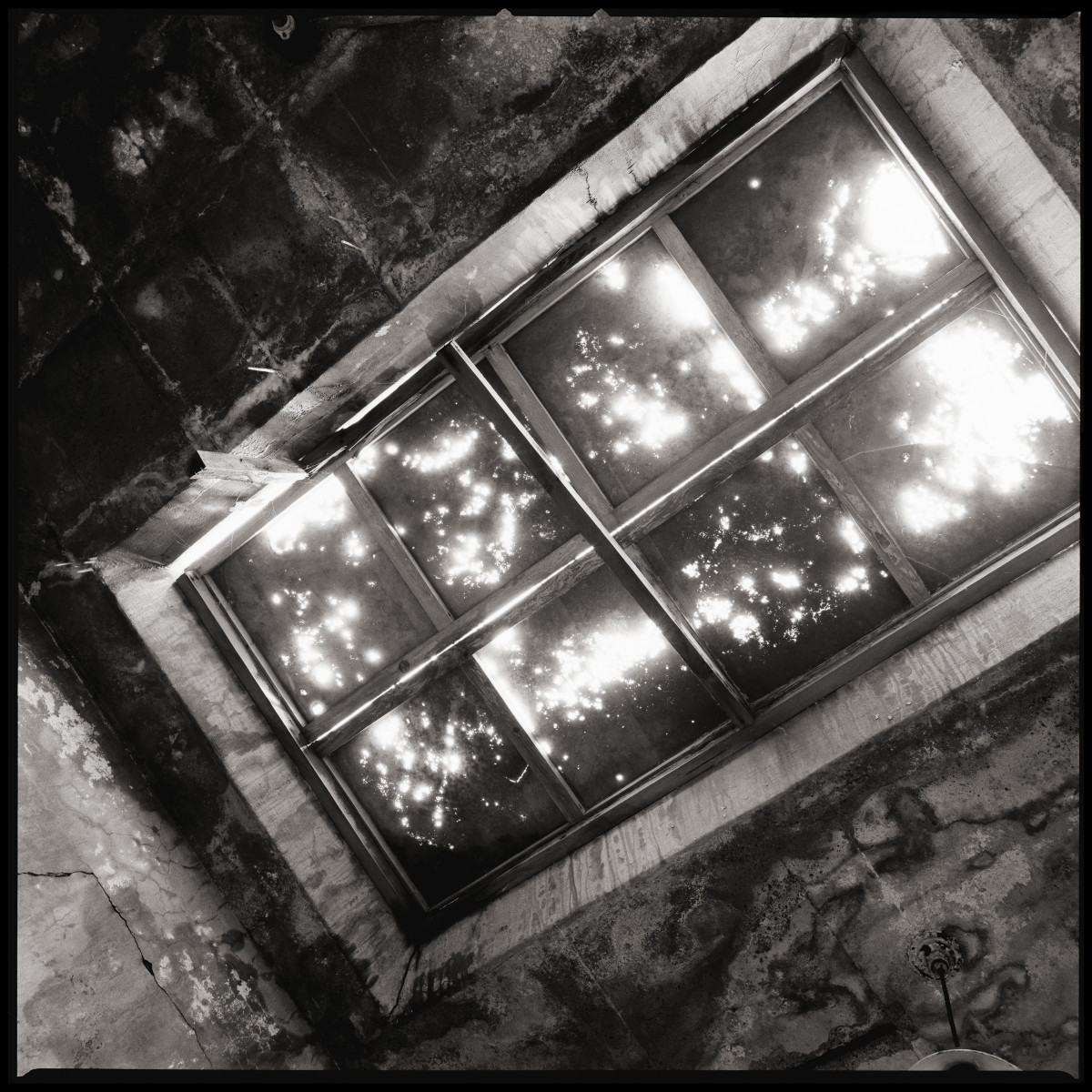 Eye of God by Eric T. Kunsman  Image: ID: A black and white image shows a window in the ceiling that has light streaming through the grime that cover most of the glass.