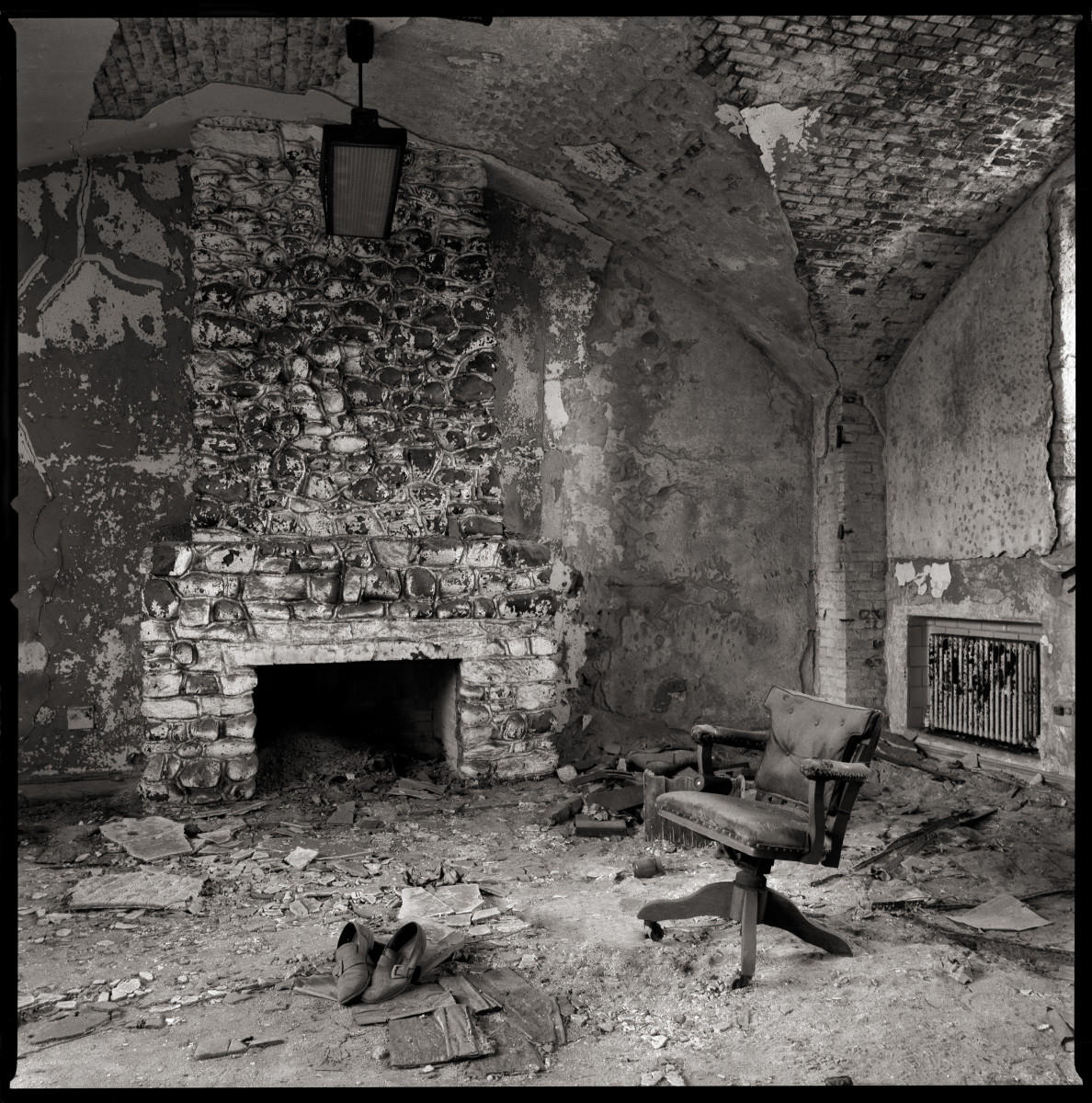 Warden's Office by Eric T. Kunsman  Image: ID: A black and white image shows a decrepit room with the back wall being an old brick fireplace and chimney.  There is a chair to the right of the fireplace.  There is debris on the ground around the chair and by the fireplace.  There is a hanging light fixture.