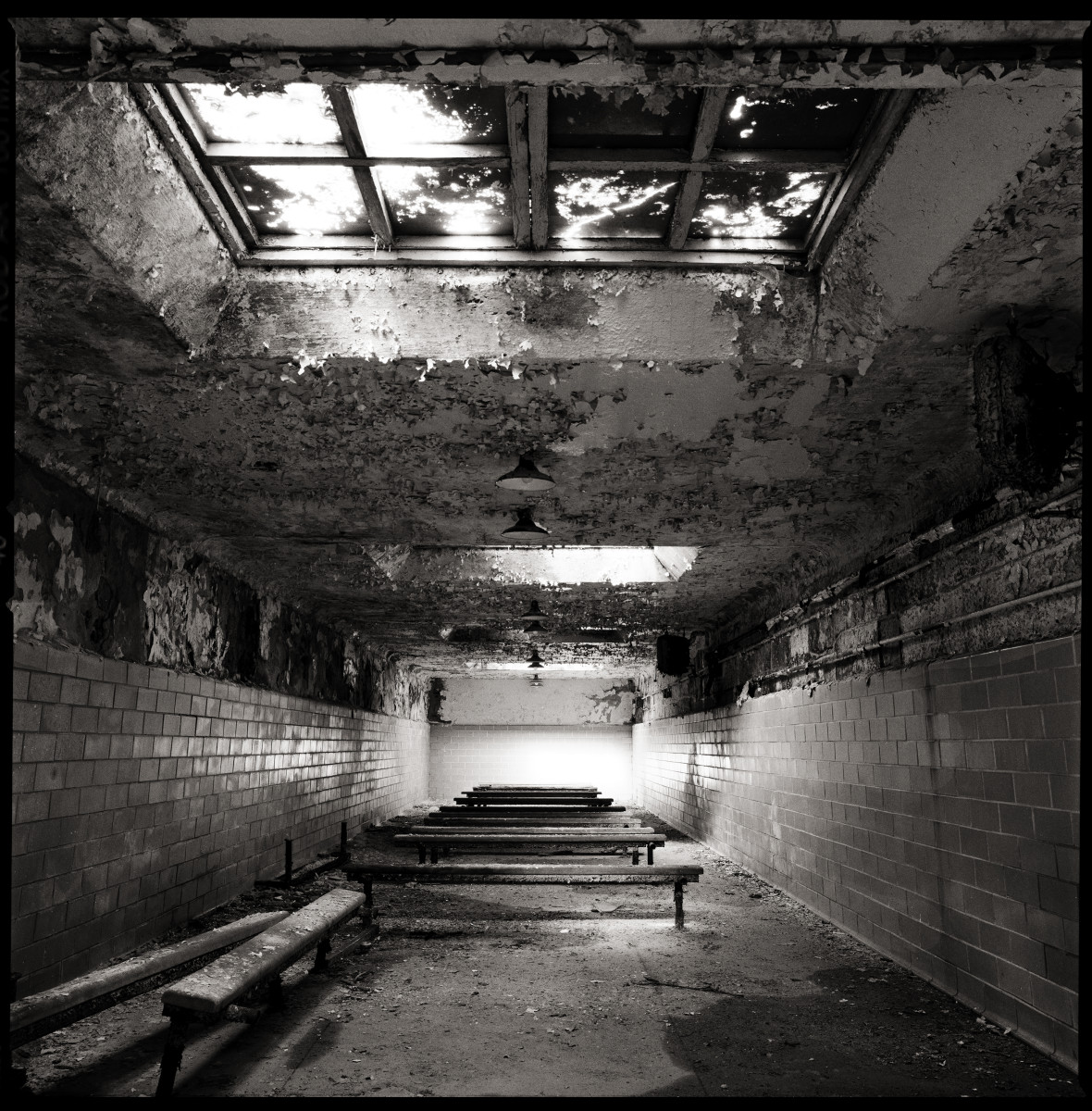 Cafeteria by Eric T. Kunsman  Image: ID: A black and white image shows a room with many benches and two large ceiling windows.  There is dust and debris built up on the floor.  The ceiling has chipping paint.