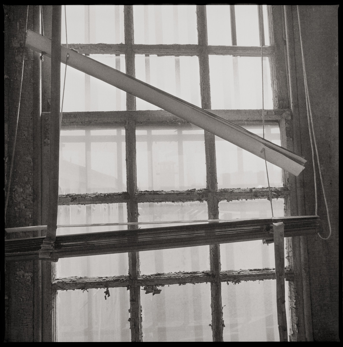 Death Row by Eric T. Kunsman  Image: ID: A black and white image shows a window with a broken shade in front of it.  The window has square pains of glass separated by wood. 