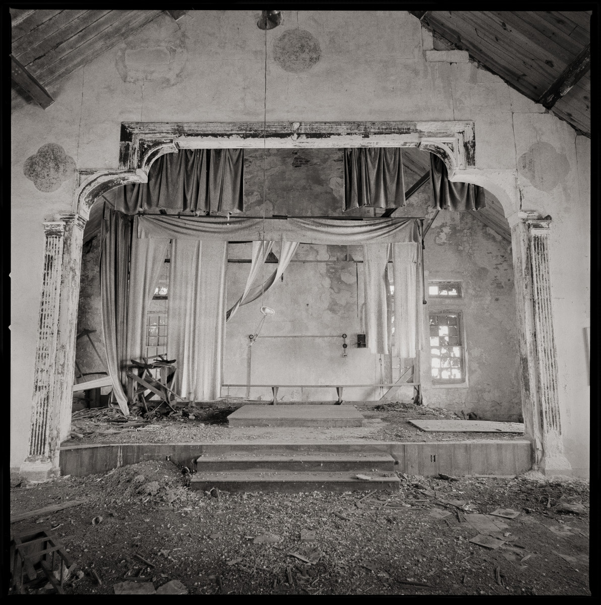 Chapel & Theater by Eric T. Kunsman  Image: ID: A black and white image shows a theater stage with curtains.  There is dirt and debris around and in front of the stage.