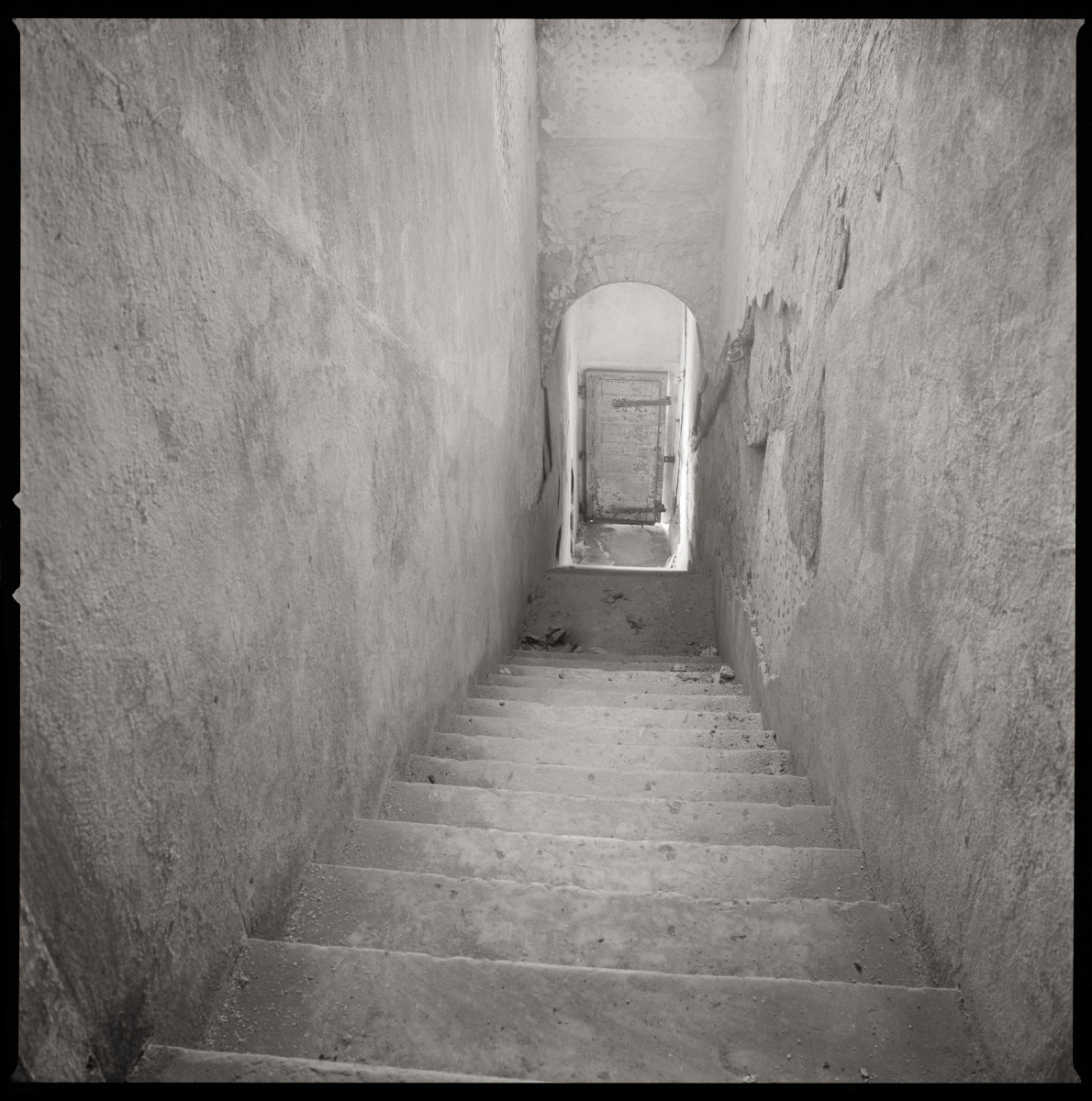 Penitence by Eric T. Kunsman  Image: ID: A black and white image shows a downwards stairway with a door at the very bottom.  The walls and stairs are cement.  