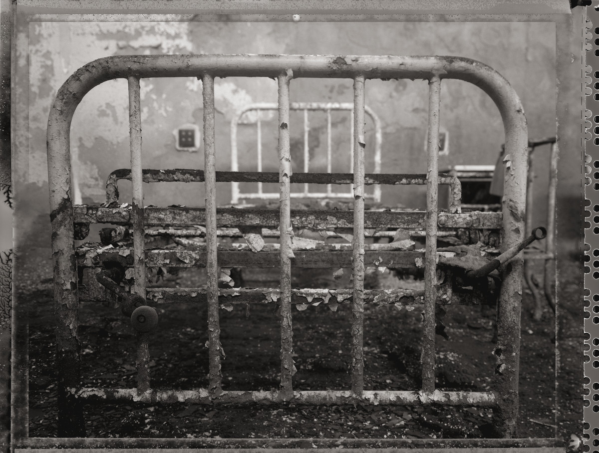 Bed, Infirmary by Eric T. Kunsman  Image: ID: A black and white image shows the foot of a metal bed frame.  The metal is rusted and the paint is chipping off.