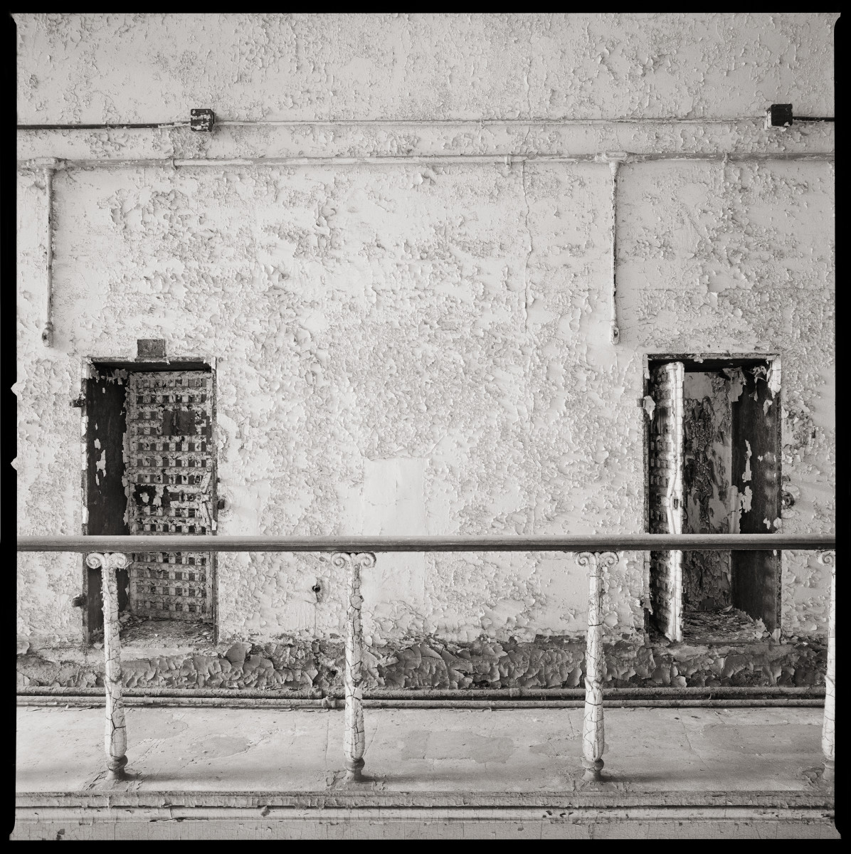 Serenity by Eric T. Kunsman  Image: ID: A black and white image shows a wall with two doors next to each other.  There is a metal bar in front of the doors.  The wall around the doors has chipping paint.