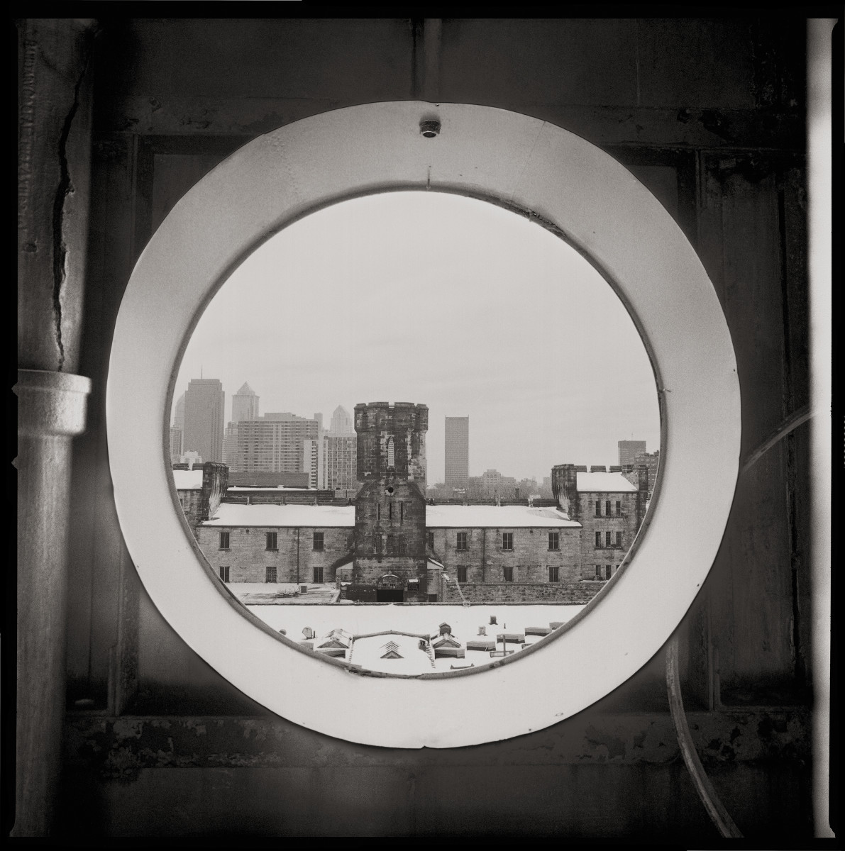 View from the Guard Tower by Eric T. Kunsman  Image: ID: A black and white image shows a view through a circular window.  A wing of the compound is visible with a tower in the center.  In the background is the Philadelphia skyline.