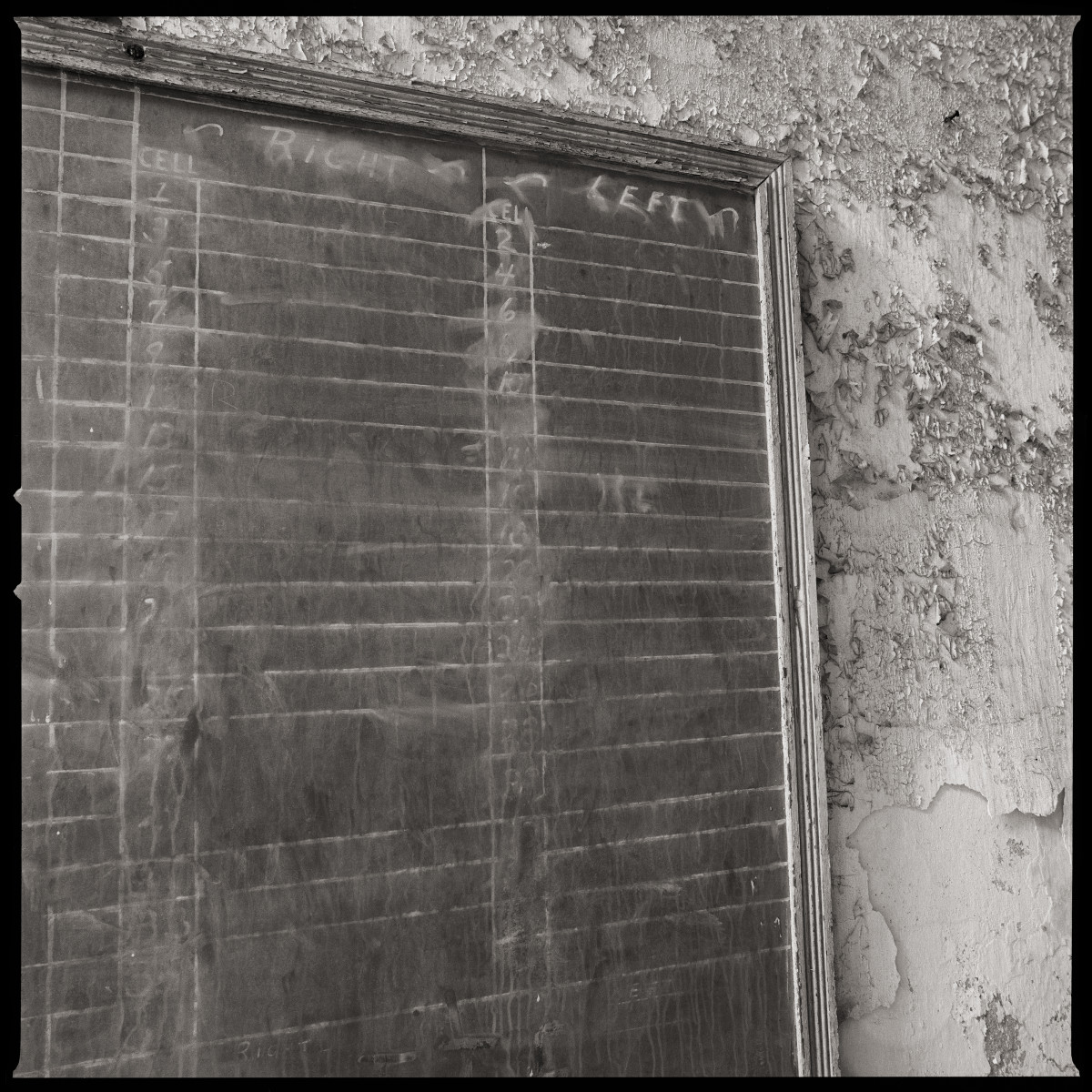 Prisoner Control Board by Eric T. Kunsman  Image: ID: A black and white image shows a chalkboard that is numbered, mounted on a wall with chipped paint,