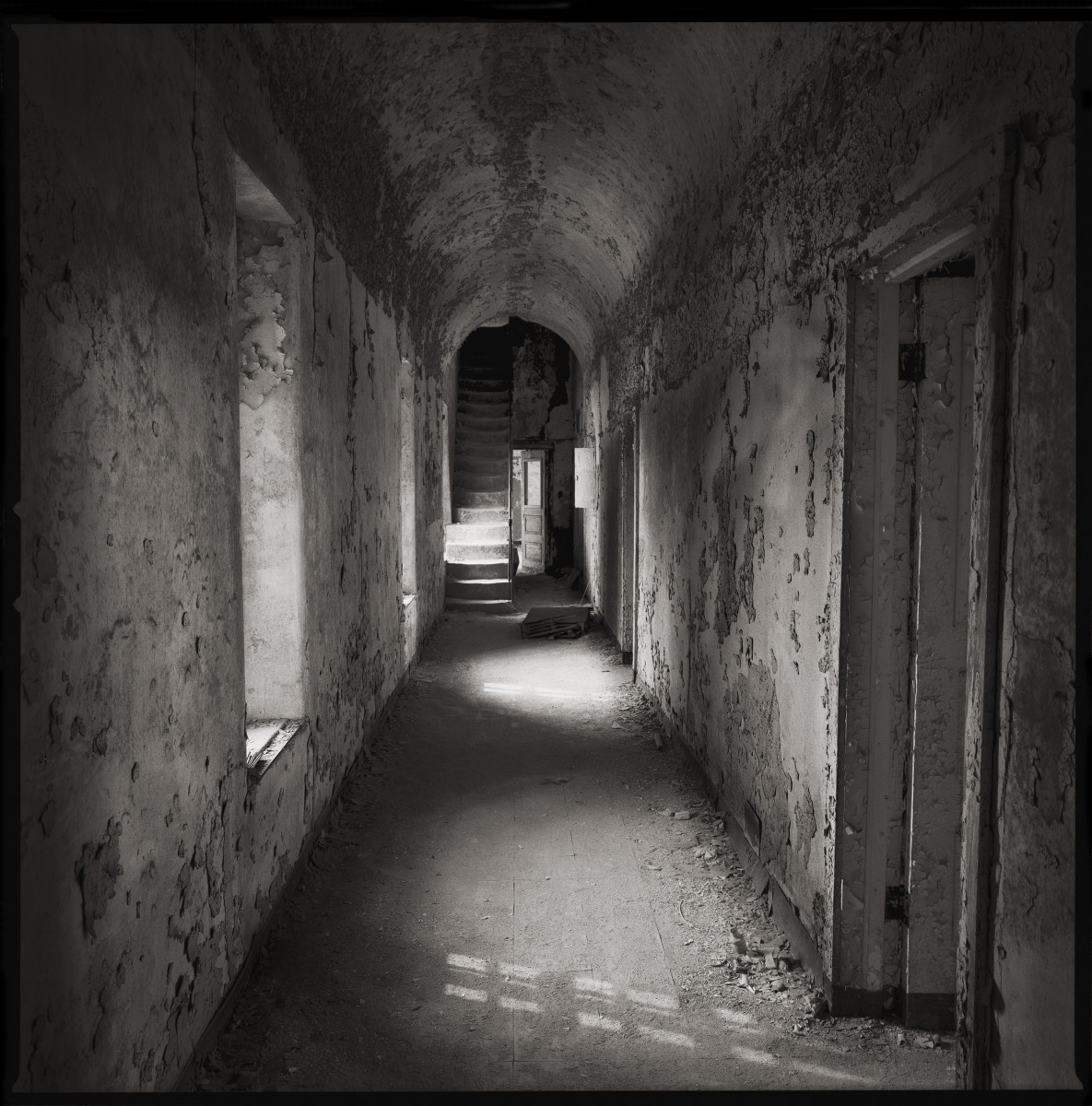 Warden's Corridor by Eric T. Kunsman  Image: ID: A black and white image shows a corridor with a curved ceiling and windows on the left wall.  There are doors on the right.  The walls and ceiling are cracking cement.