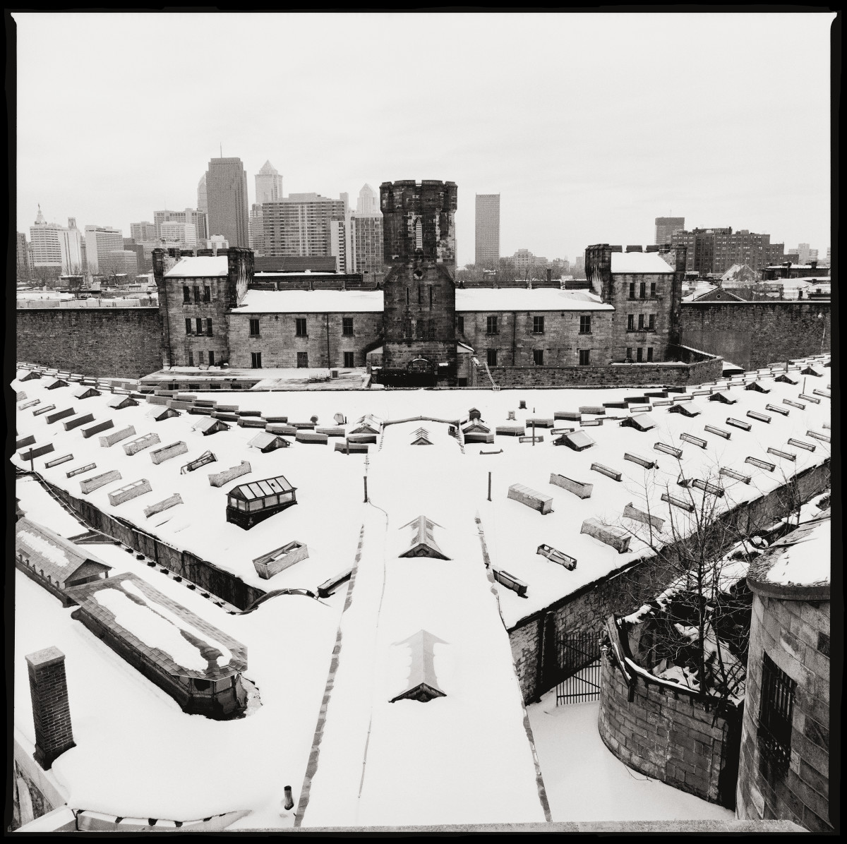 Looking Towards Downtown Philadelphia by Eric T. Kunsman  Image: ID: A black and white image shows a view of the compound with the Philadelphia skyline in the background.  The compound has a wing in the center, and two branching off to each side from the center.  There is also a wing in the back.  The image is snowy and grey.