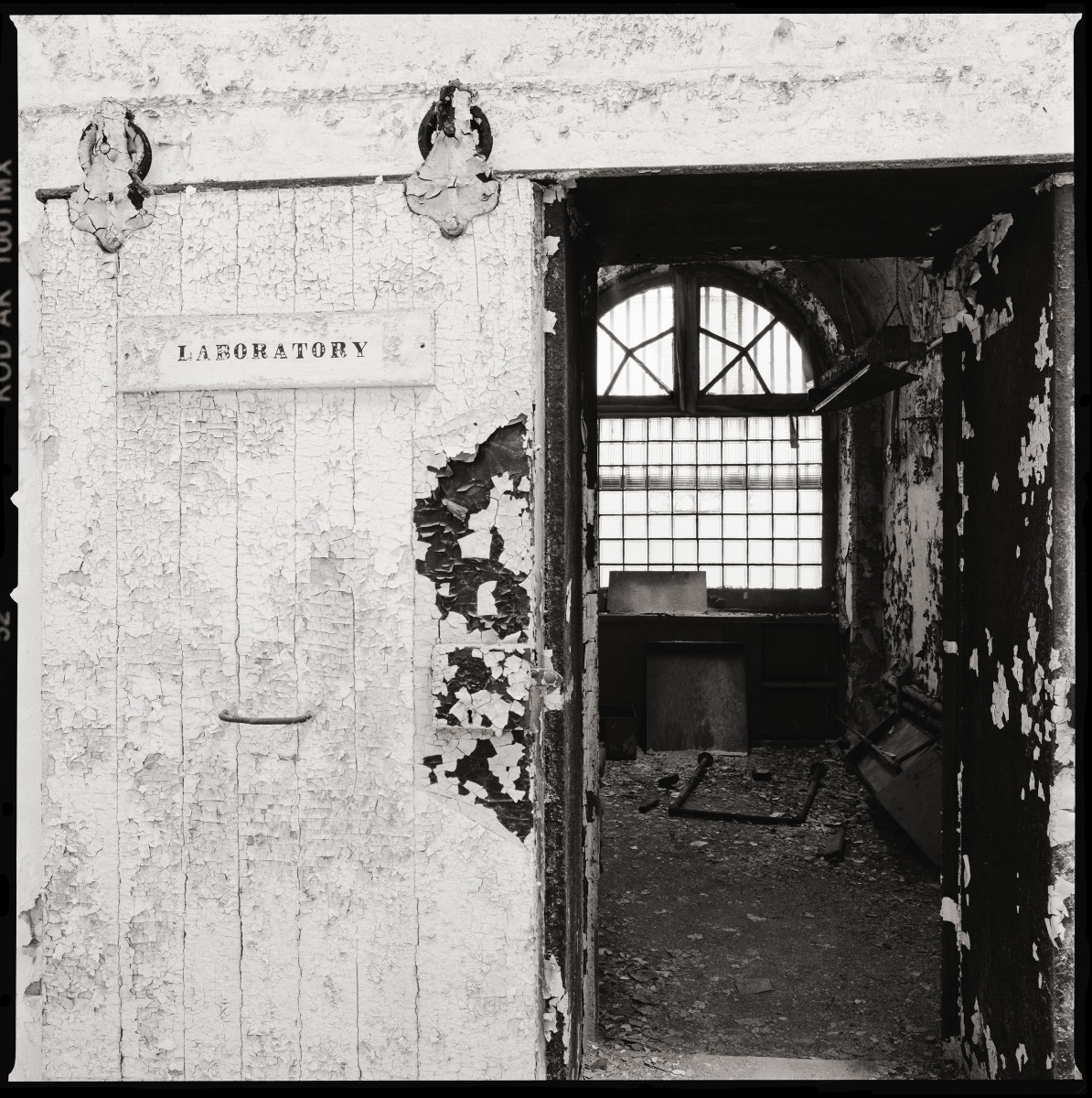 Laboratory by Eric T. Kunsman  Image: ID: A black and white image shows an open doorway entrance with a sign for "Laboratory" on the left on the door.  On the right the room is visible.  There is a large window with bars across it.  There is a lot of dirt and debris on the ground in the room.