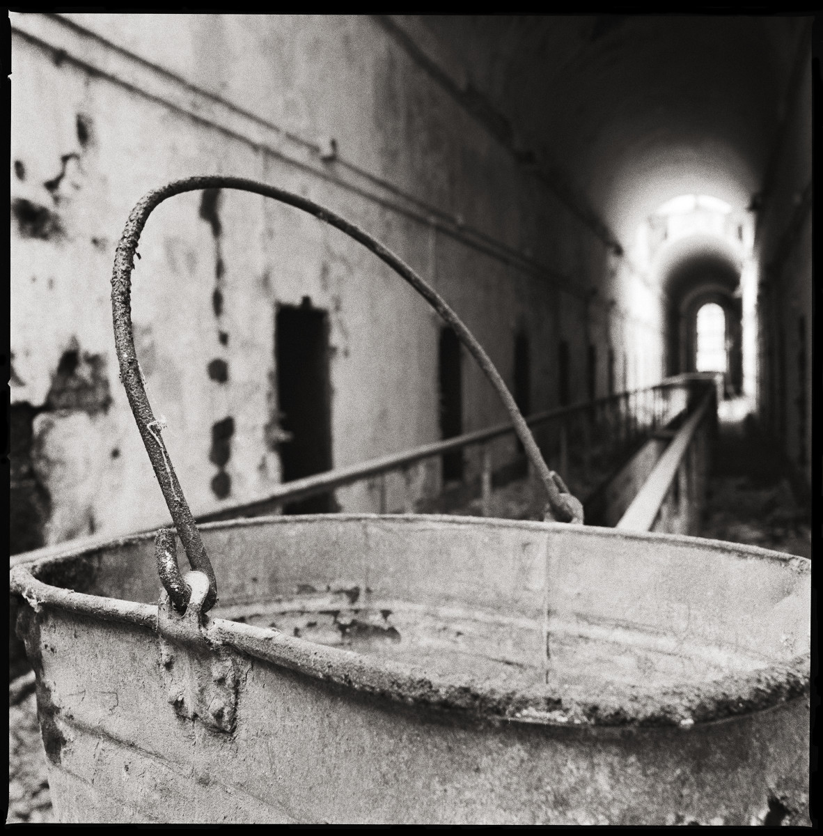Bucket, Cellblock #6 by Eric T. Kunsman  Image: ID: A black and white image shows a rusted metal bucket in the foreground. In the background, prison cells are visible but not in focus.