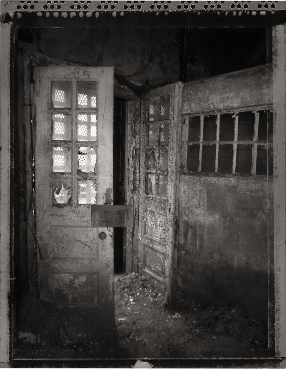 Entrance to the Infirmary by Eric T. Kunsman  Image: ID: A black and white image shows a doorway that is open. The room inside is not visible.  The doors have windows in them and are grimy.  The paint is cracking and there are piles of dirty on the floor.