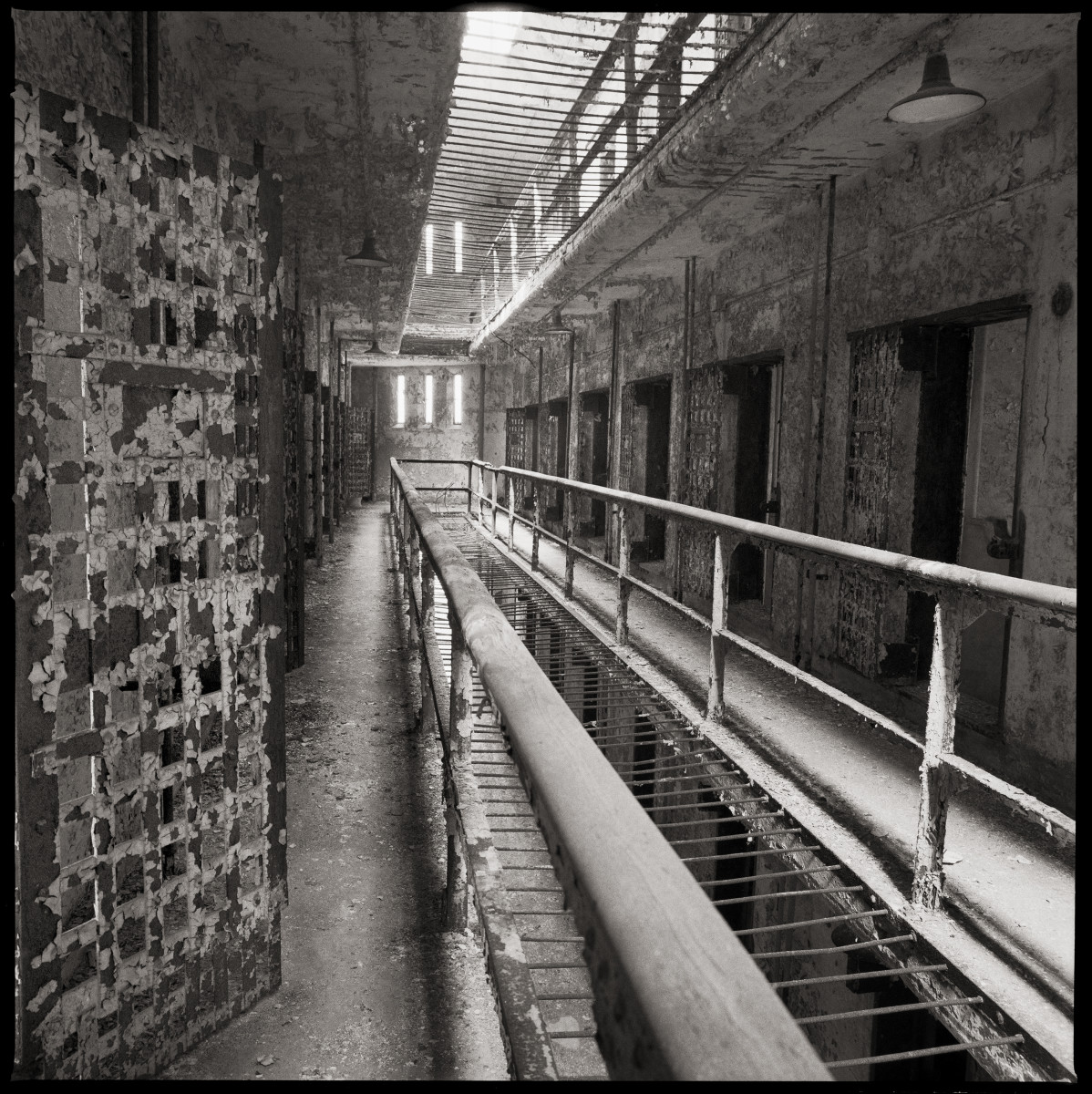 Cellblock #14 by Eric T. Kunsman  Image: ID: A black and white image shows a corridor of prison cells.  There is a hallway on either side of the open center with cells on both sides.  There is a window at the end of the corridor.