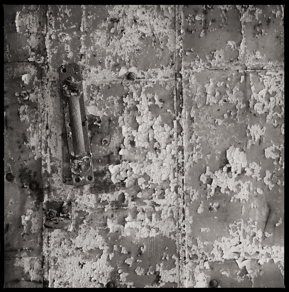 Untitled by Eric T. Kunsman  Image:  ID: A black and white image shows a close up of a handle on the wall.  There is an abundance of grime on and around the handle.  The paint on the wall is peeling.  The handle is vertical, but has been shifted right over the years.