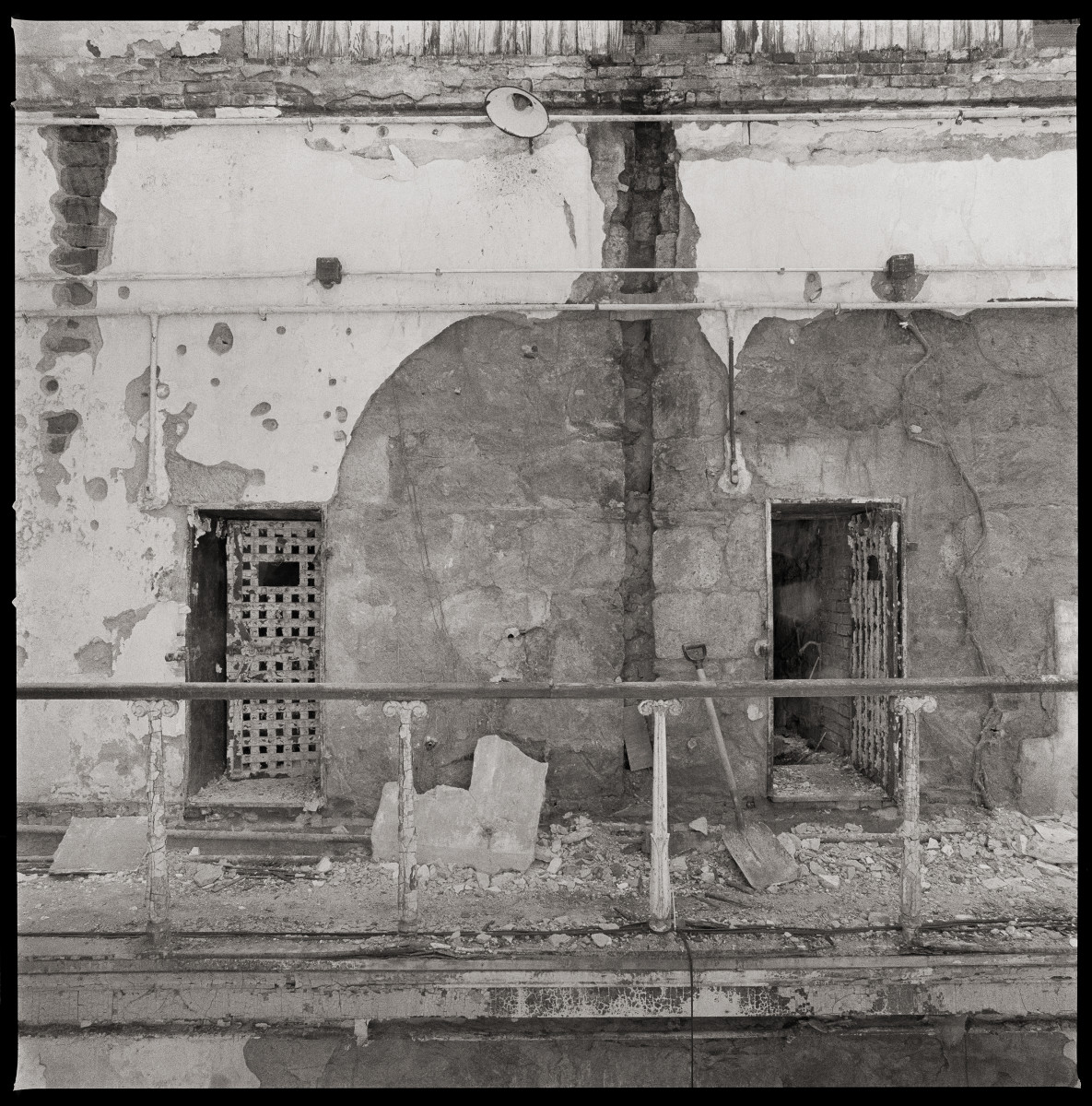 Untitled by Eric T. Kunsman  Image: ID: A black and white image shows two cells side by side from across the partition. There is a bar in front of the hall.  The door on the left is closed; the one on the right is open.  Around the doors is a wall of cracking cement.