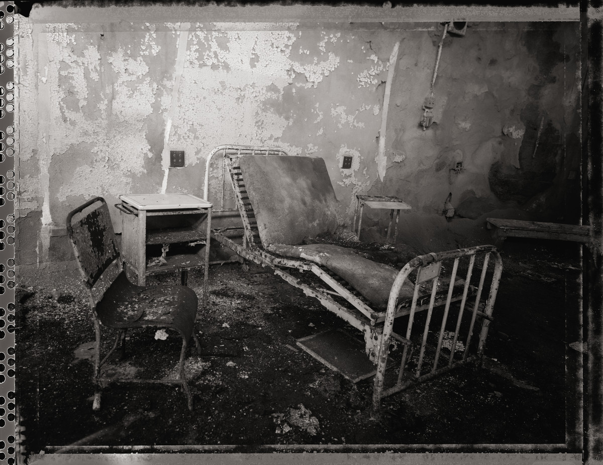 Waiting in the Infirmary by Eric T. Kunsman  Image: ID: A black and white image shows a room with a bent metal bed in the center.  To the left of the bed is a chair and a side table.  The room is dirty and the paint is peeling off the walls.