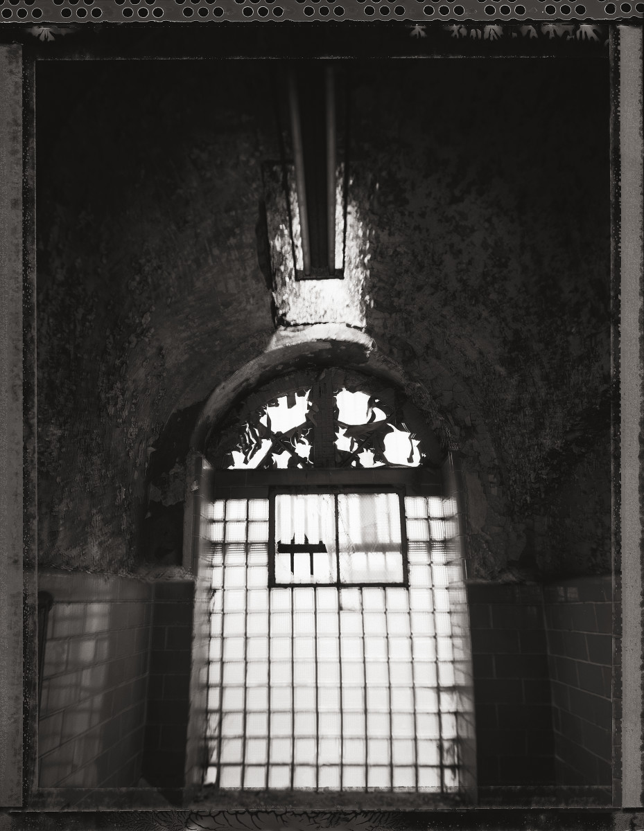 Infirmary by Eric T. Kunsman  Image: ID: A black and white image shows a large glass window with metal bars on it.  There is a rectangular light hanging from the ceiling. 