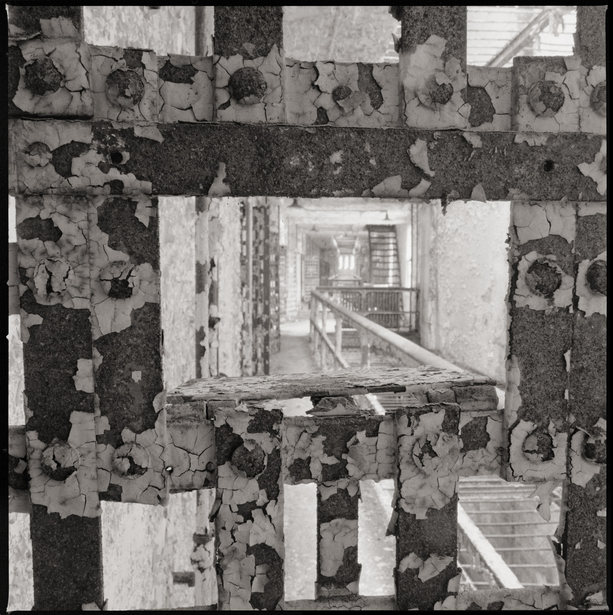 Cellblock #14 by Eric T. Kunsman  Image: ID: A black and white image shows a view of the cellblock from inside a cell, through the metal door.  There is a railing to the right and more cells to the left.