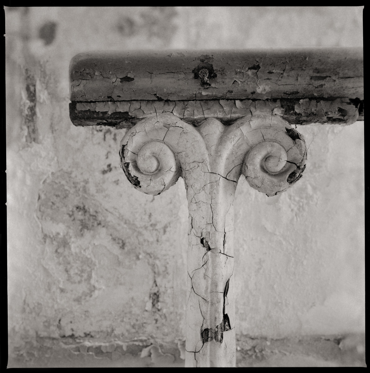 Detail of a Railing by Eric T. Kunsman  Image: ID: A black and white image shows a detail of a railing.  The top is flat wood, but the metal pole supporting it had two curls on each side.  The metal is painted white but it is chipping.