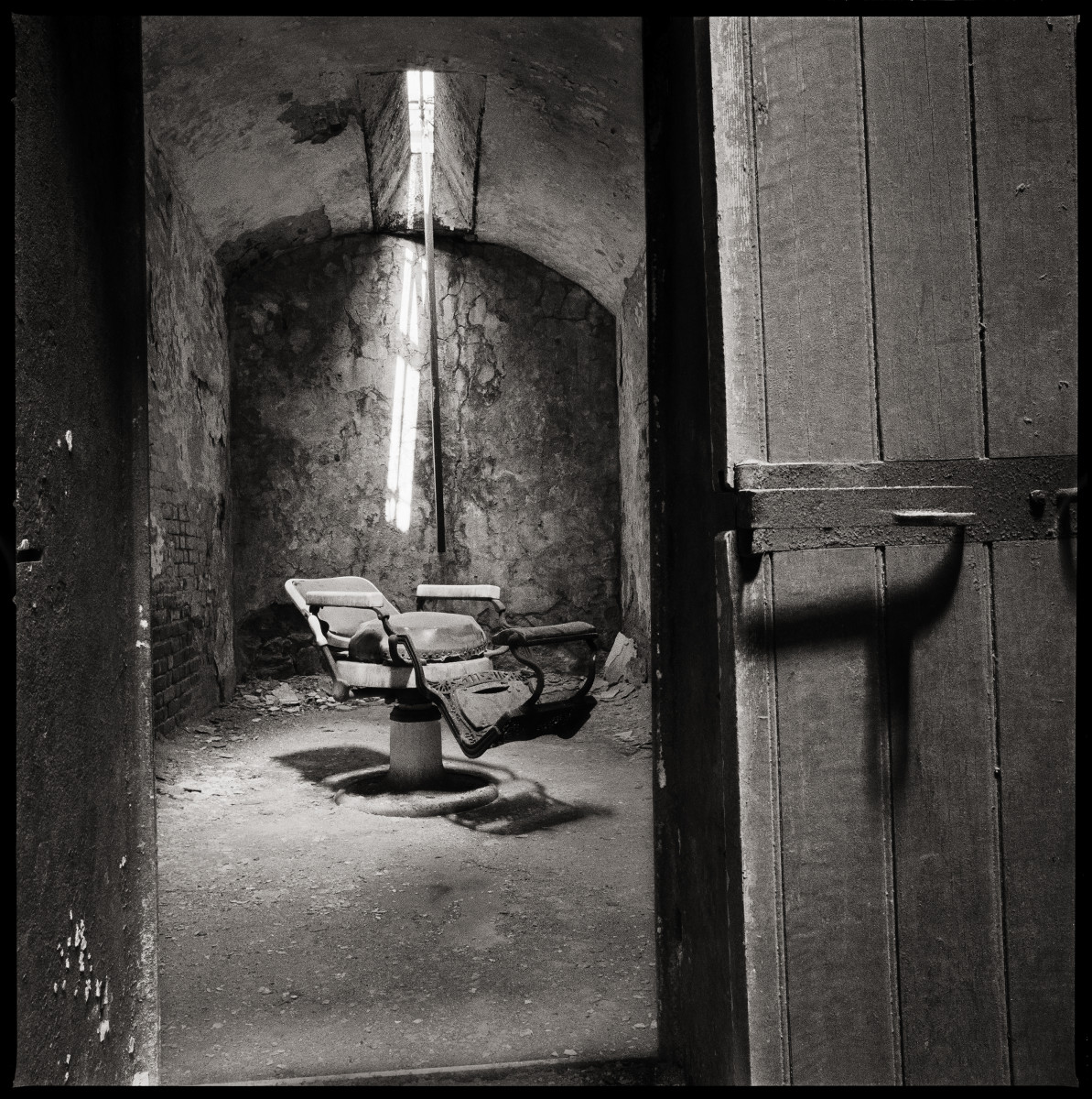 Dentist Chair by Eric T. Kunsman  Image: ID: A black and white image shows a room with an open door.  Inside the room there is an old dentist chair with a beam of light on the back wall.