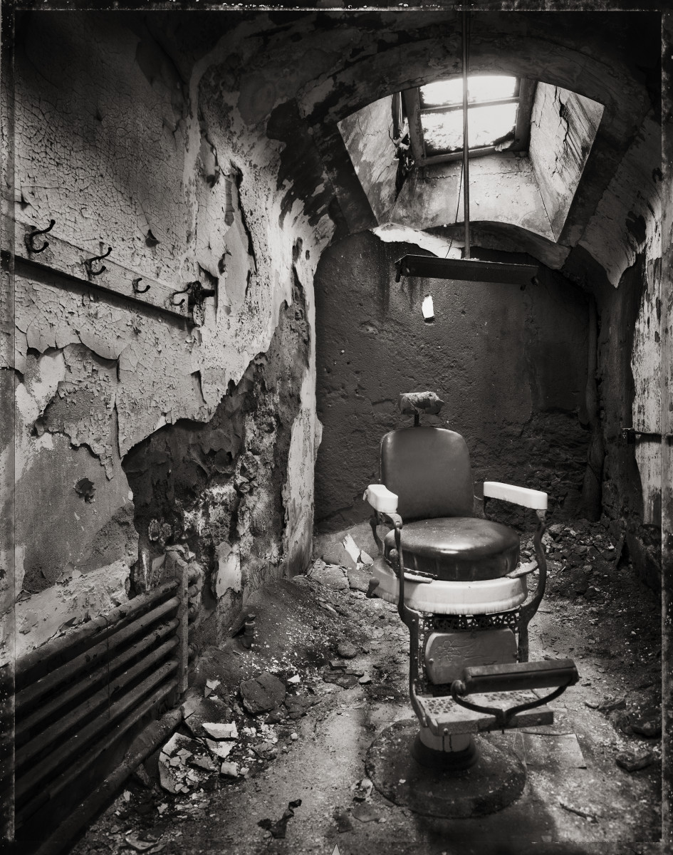 Barber Chair by Eric T. Kunsman  Image: ID: A black and white image shows a room with a large ceiling window in the curved ceiling.  The walls and ceiling have cracked plaster covering now-exposed brick. There is a barber chair in the middle of the room and coat hooks on the left wall.