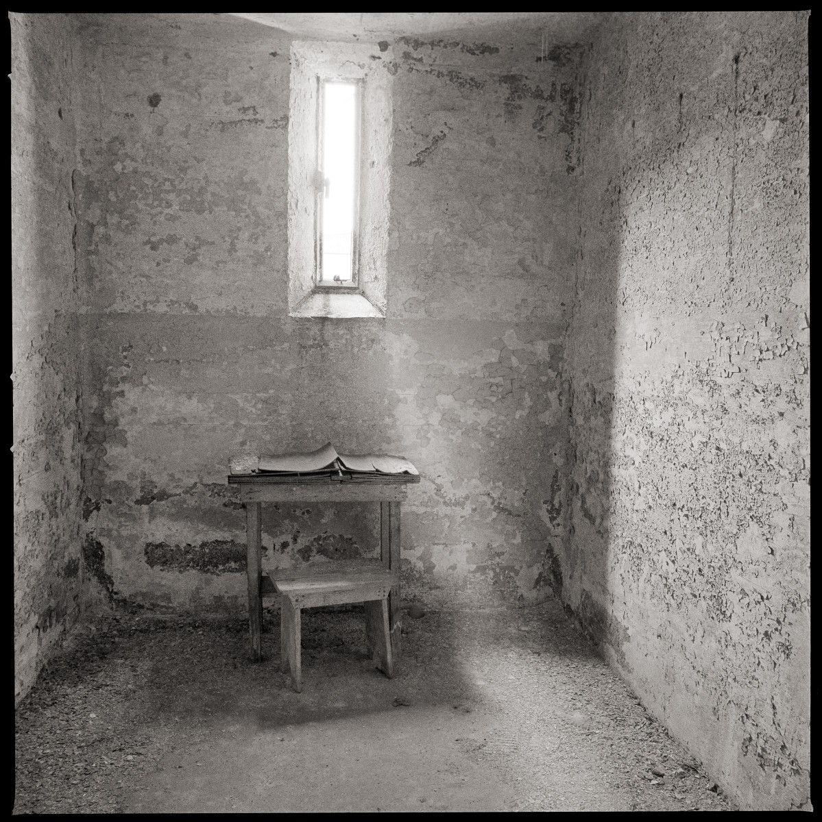 Divine Intervention by Eric T. Kunsman  Image: ID: A black and white image shows a desk below a window in an otherwise empty room.  The paint on the wall is cracked.  There is a beam of light coming through the window.