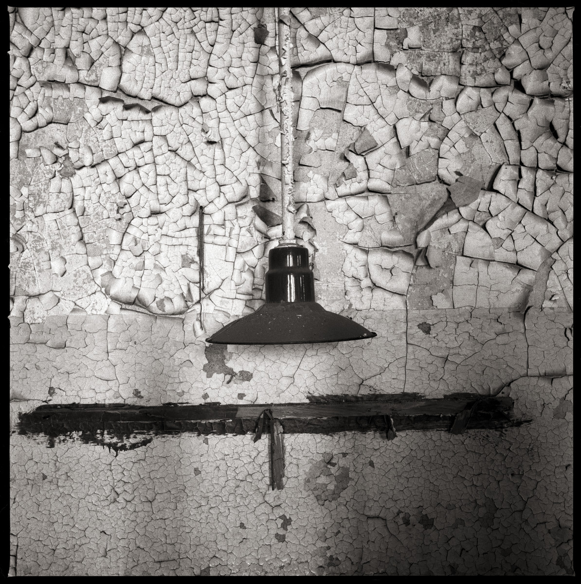 Kitchen by Eric T. Kunsman  Image: ID: A black and white image shows a close up of a hanging light fixture next to a wall. The fixture has a circular bottom and it attached to a pole from the ceiling.  The wall has cracking paint and cement.