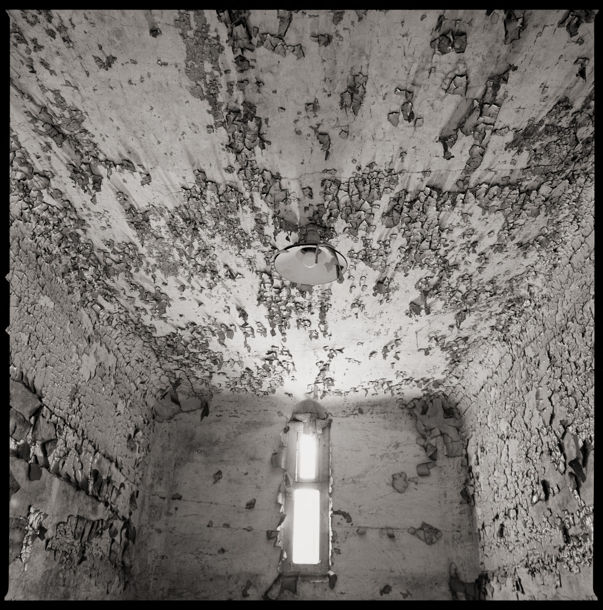 Untitled by Eric T. Kunsman  Image: ID: A black and white image shows an upward view of a room.  The walls and ceiling are cracked cement with peeling paint.  The back wall has a narrow window with a curved top.