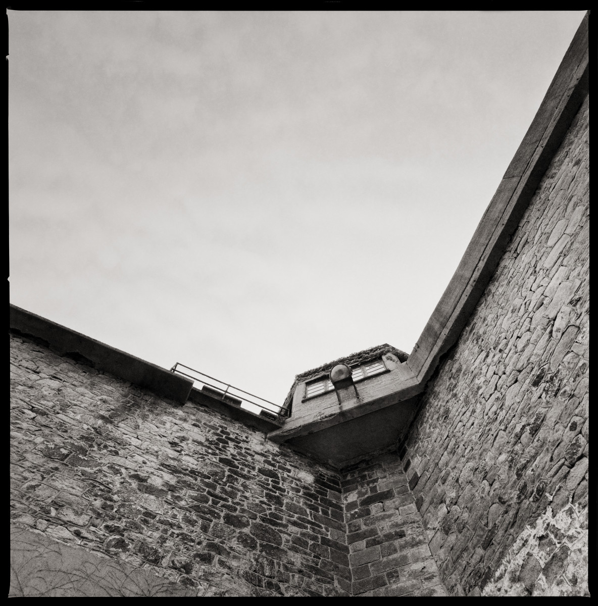 Guard Tower by Eric T. Kunsman  Image: ID: A black and white image shows a upwards view of the corner guard tower.  There are not clouds in the sky.  The tower has walls that extend from the corner.  The building is brick.