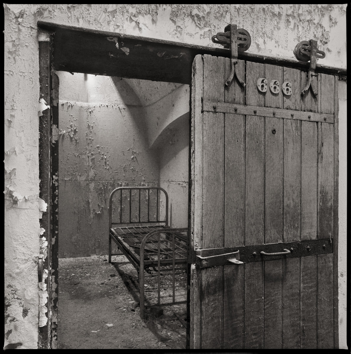 Devil has their Soul by Eric T. Kunsman  Image: ID: A black and white image shows room number 666 with ht door ajar.  There is a metal bed frame inside of the room.  The door is wooden with a metal bar in the center.