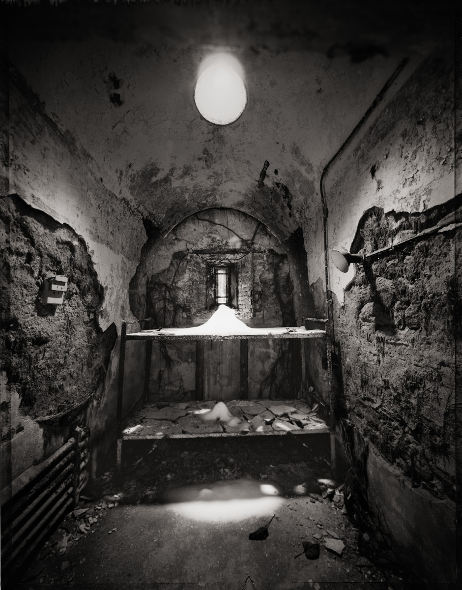 Bunkmates by Eric T. Kunsman  Image: ID: A black and white image shows a room with a curves ceiling and the plaster coming off the walls.  Against the back wall there is a metal bunkbed frame.  There is a pile of dust on the top bunk and starting on the bottom bunk.  There is a light fixture on the ceiling in the middle of the room and a small window on the back wall.