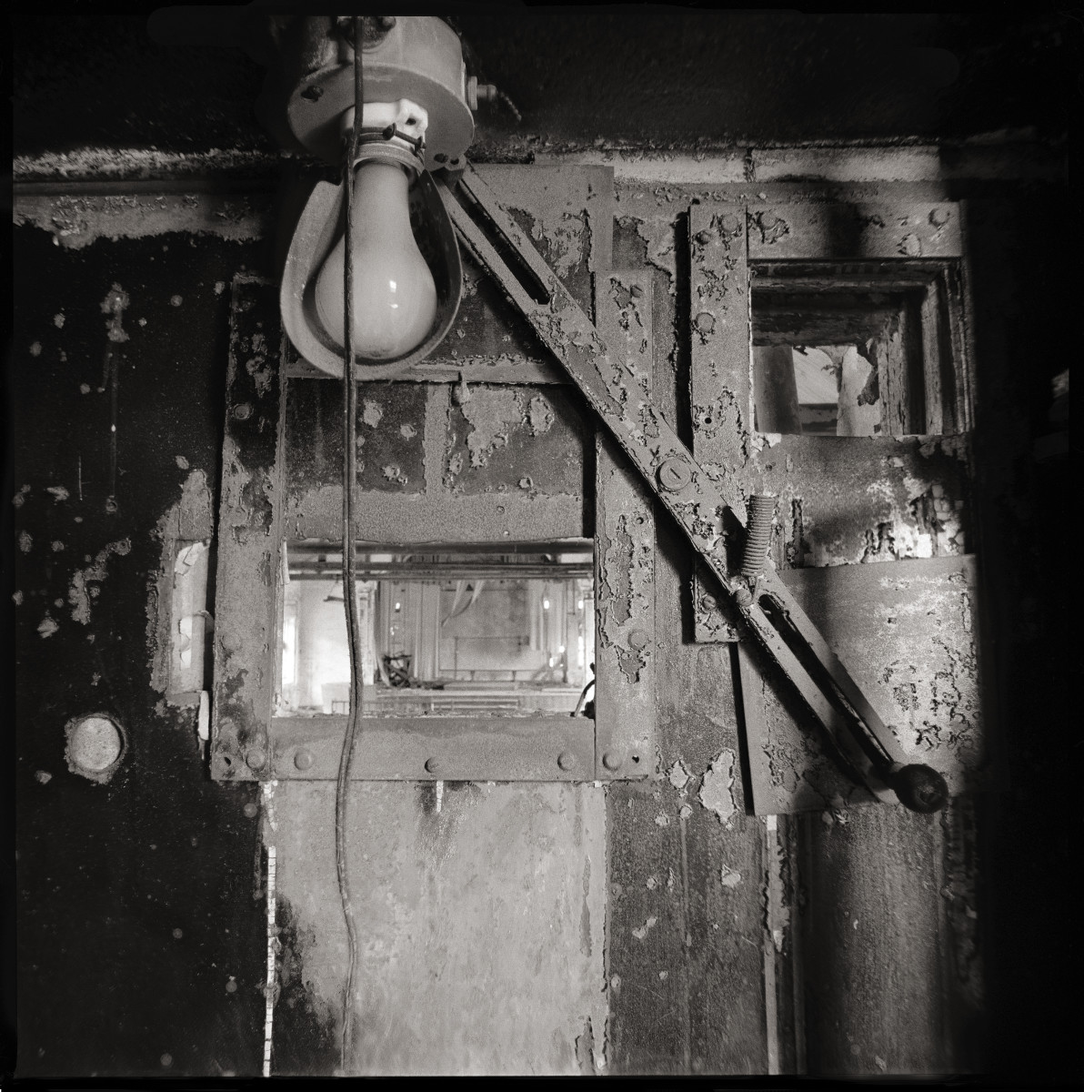 Projection Room by Eric T. Kunsman  Image: ID: A black and white image shows a lightbulb by a small glass window into the next room where projections were displayed.