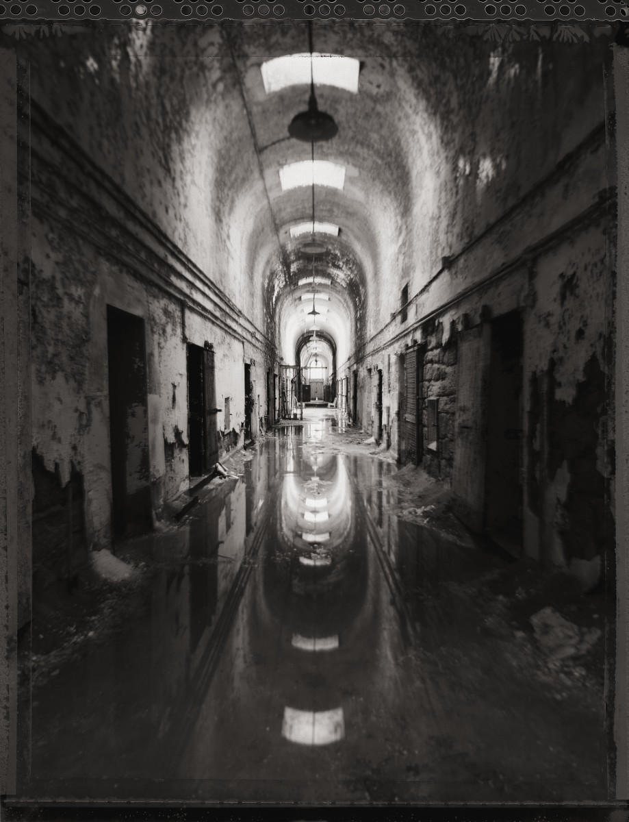Infirmary by Eric T. Kunsman  Image: ID: A black and white image shows a corridor with a high curved ceiling.  There are light fixtures hanging from the ceiling as well as windows in the ceiling.  The corridor is reflected onto the floor due to it having standing water.