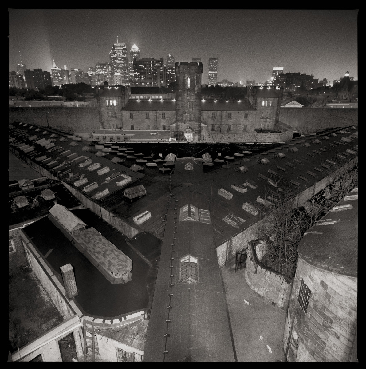 Looking Towards Downtown Philadelphia by Eric T. Kunsman  Image: ID: A black and white image shows a view of the compound with the Philadelphia skyline in the background.  The compound has a wing in the center, and two branching off to each side from the center.  There is also a wing in the back.  The image was taken at night, and the lights from downtown Philadelphia are twinkling.